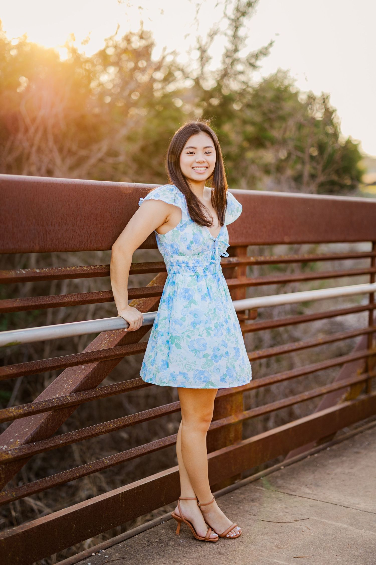 Young woman in blue floral dress posing on rustic bridge at golden hour sunset.