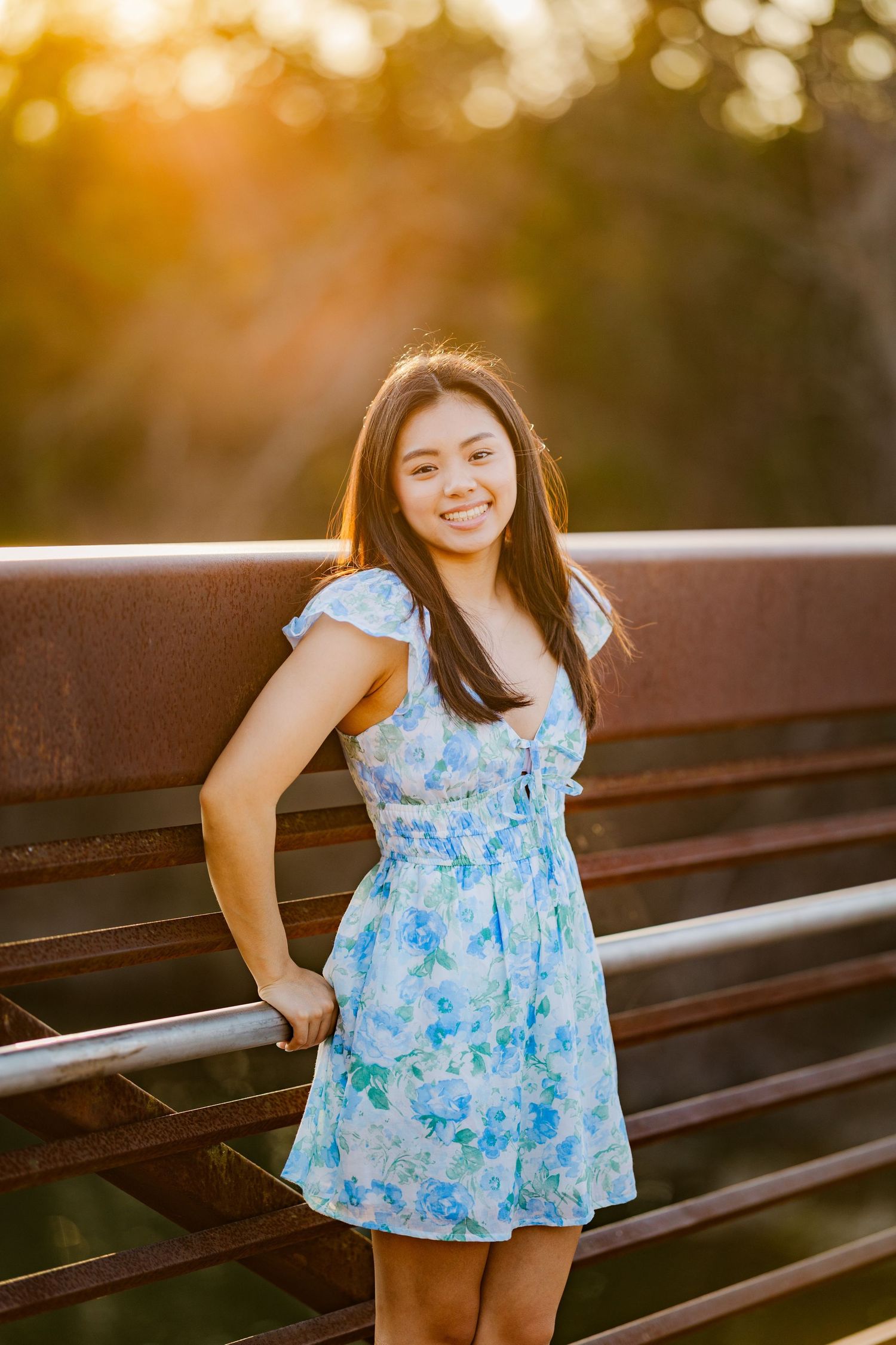 Smiling young woman in blue floral dress leaning on rustic bridge railing at golden hour.