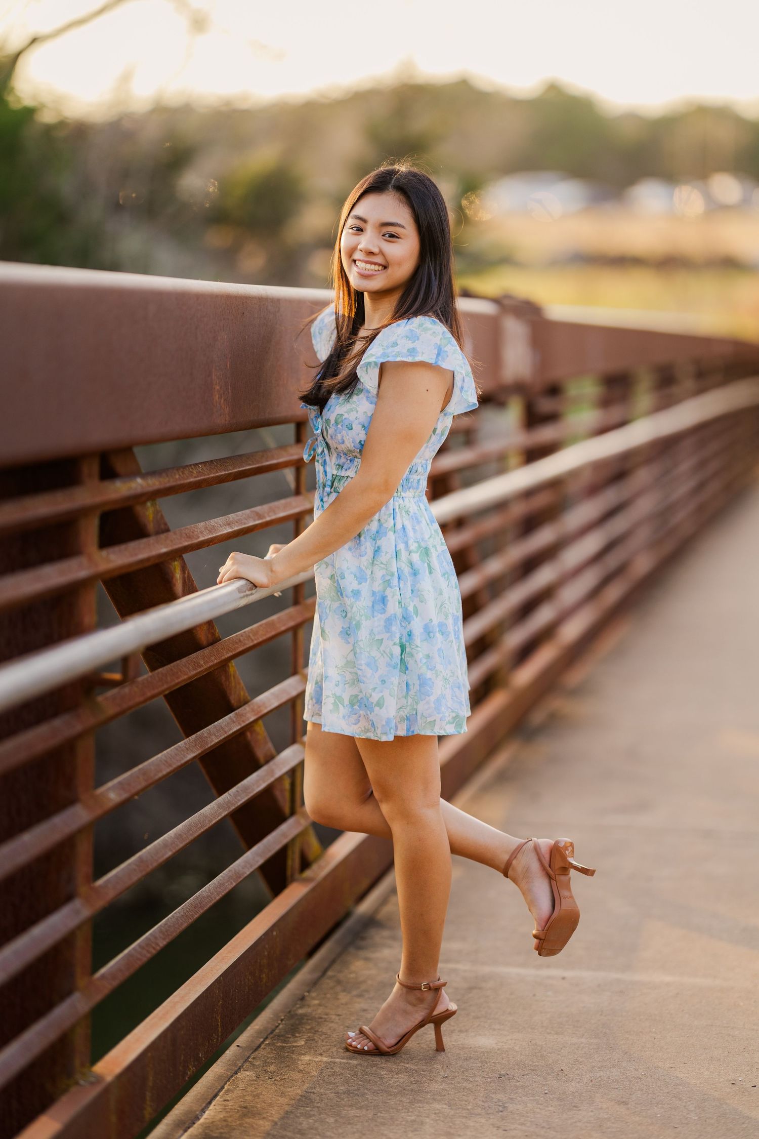 Smiling teen girl in blue floral dress poses on a rustic bridge railing at golden hour sunset.