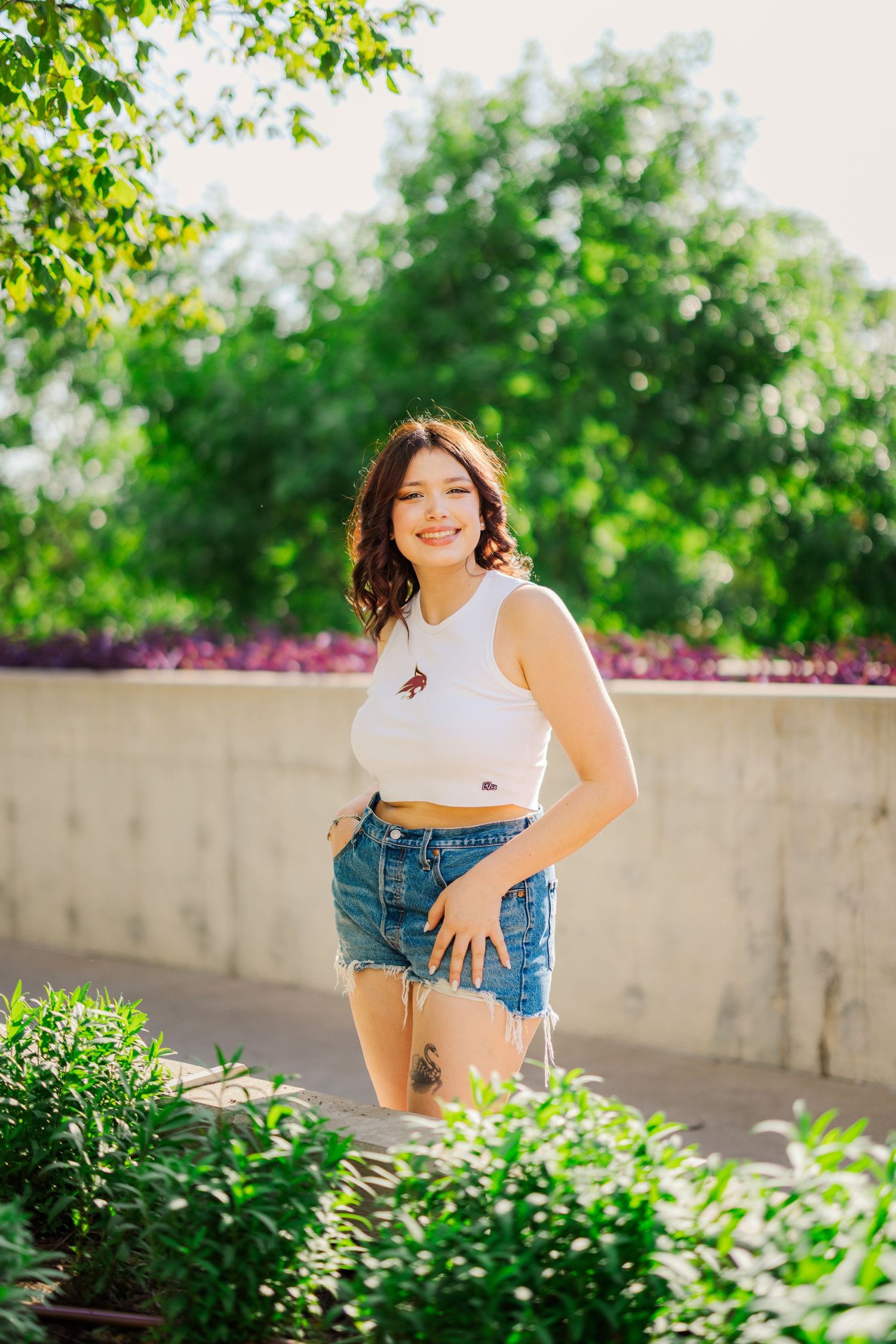 Smiling young woman in white crop top and denim shorts standing among green plants outdoors.