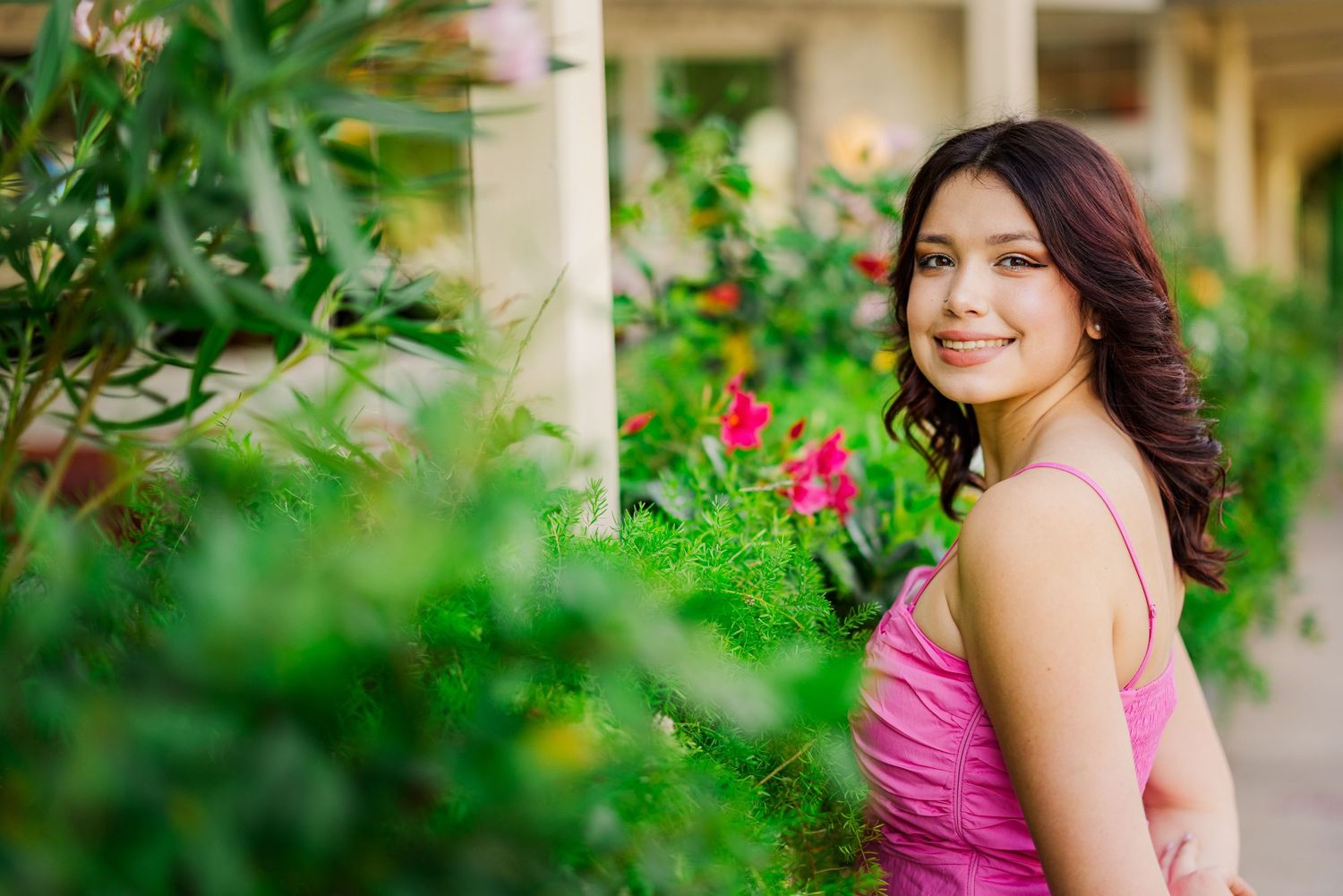 Smiling young woman in pink dress posing among colorful garden flowers and green foliage.