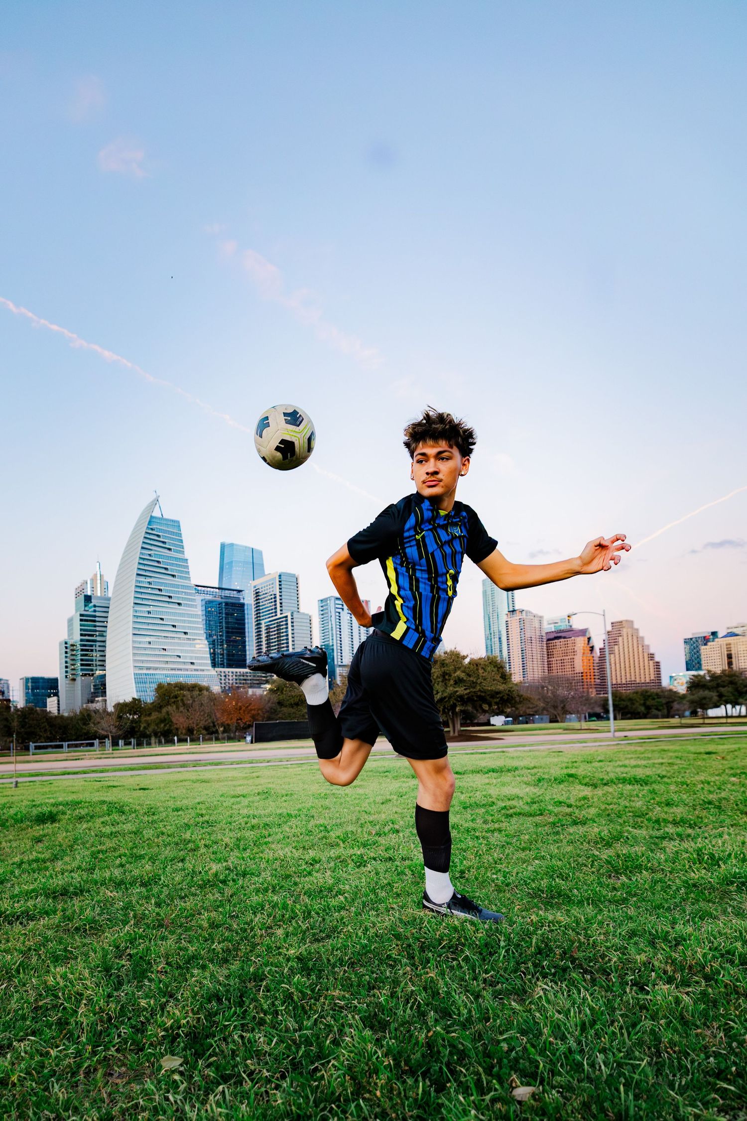 Athletic teen in soccer uniform skillfully juggles ball on green field with city skyline at sunset.