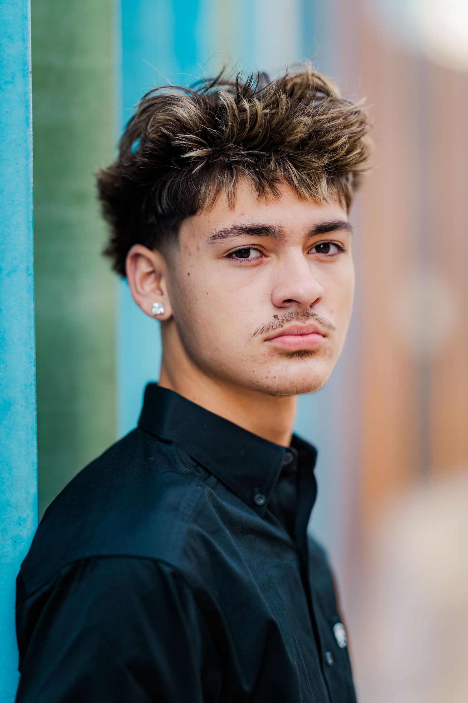 Teen boy in black shirt leans against colorful teal wall with serious expression wearing small stud earring.