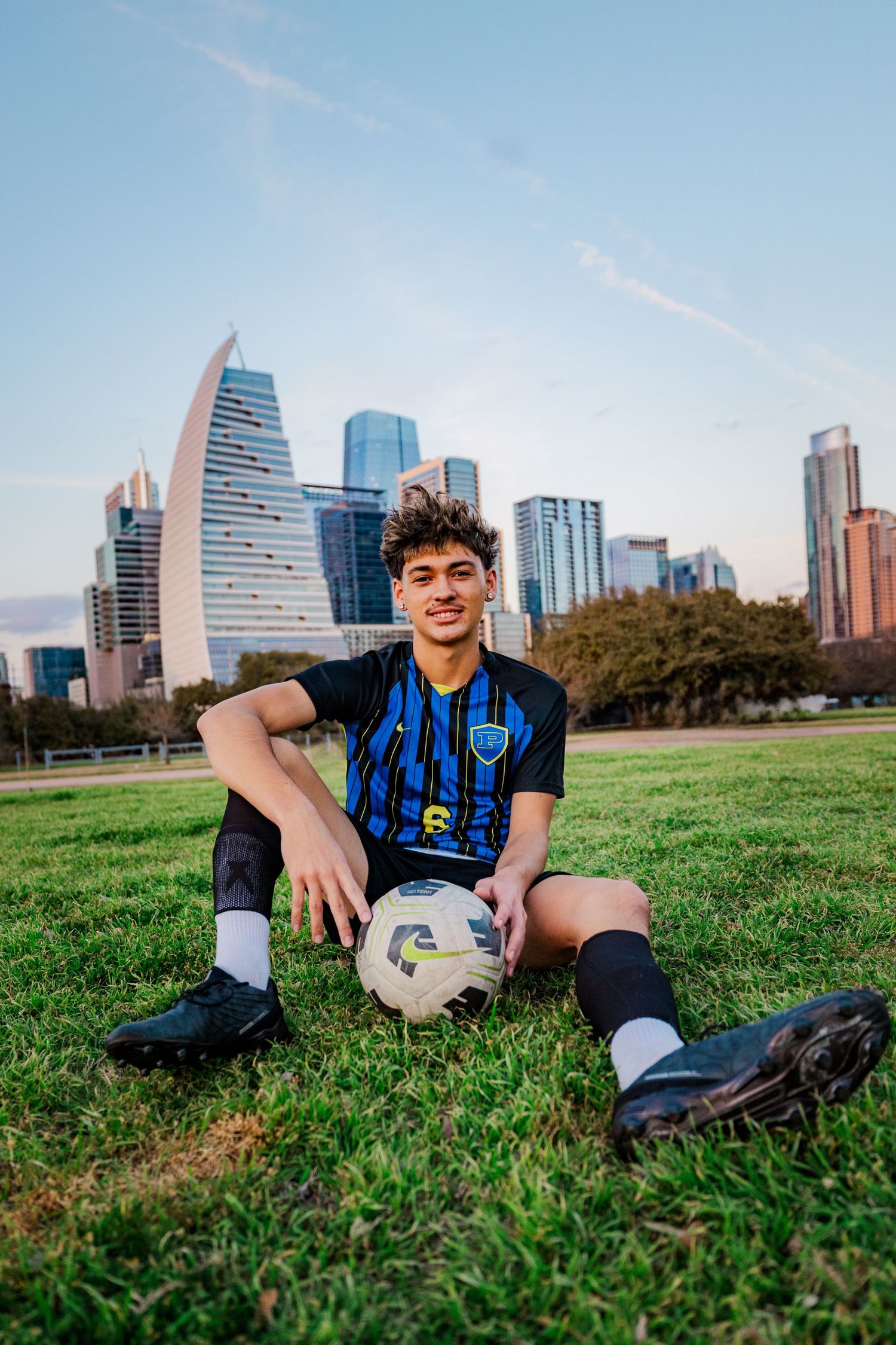Teen soccer player in blue and black jersey sits on grass with ball against city skyline backdrop.