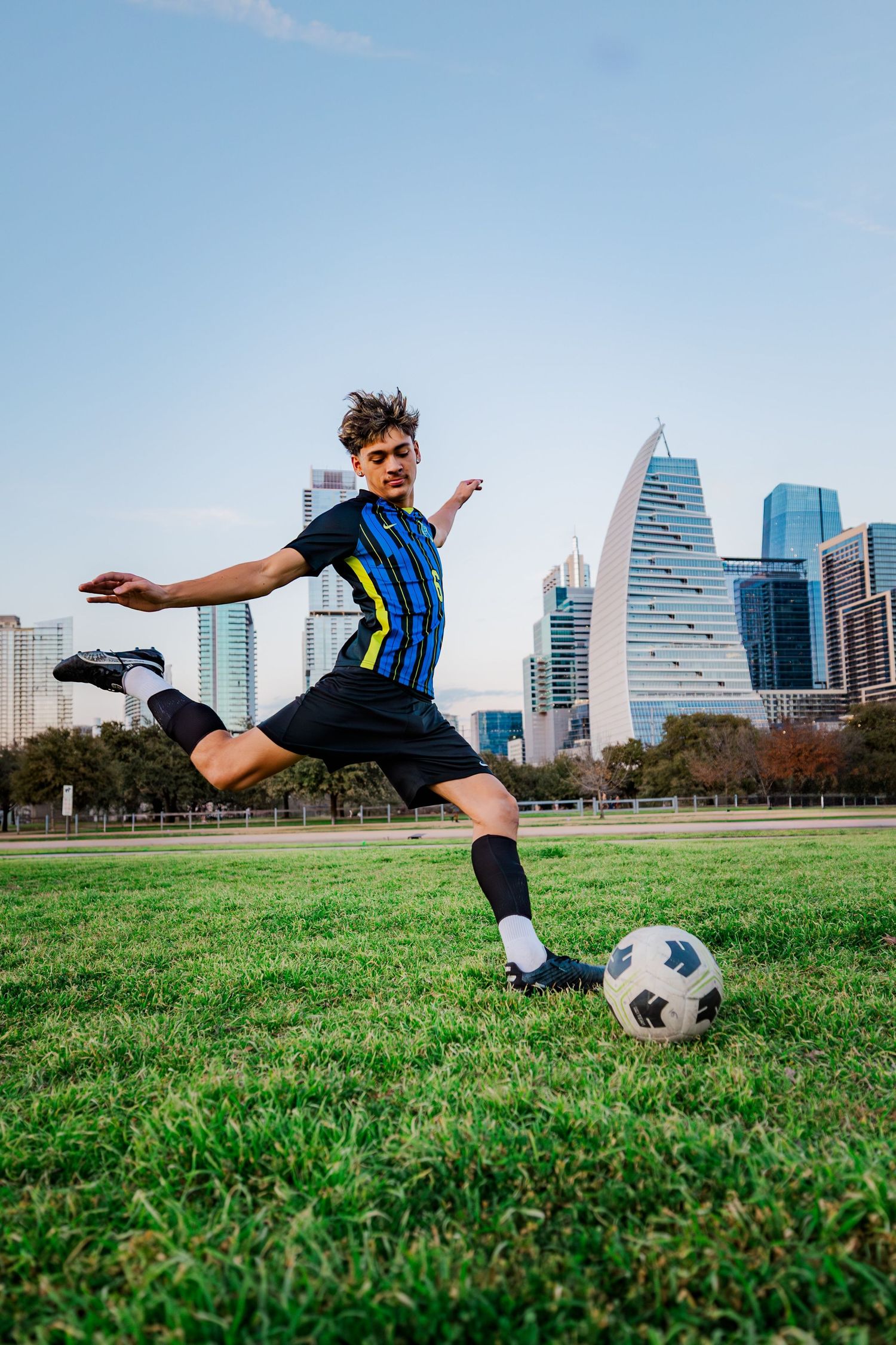 Young male soccer player kicking a ball on grass with city skyline backdrop.