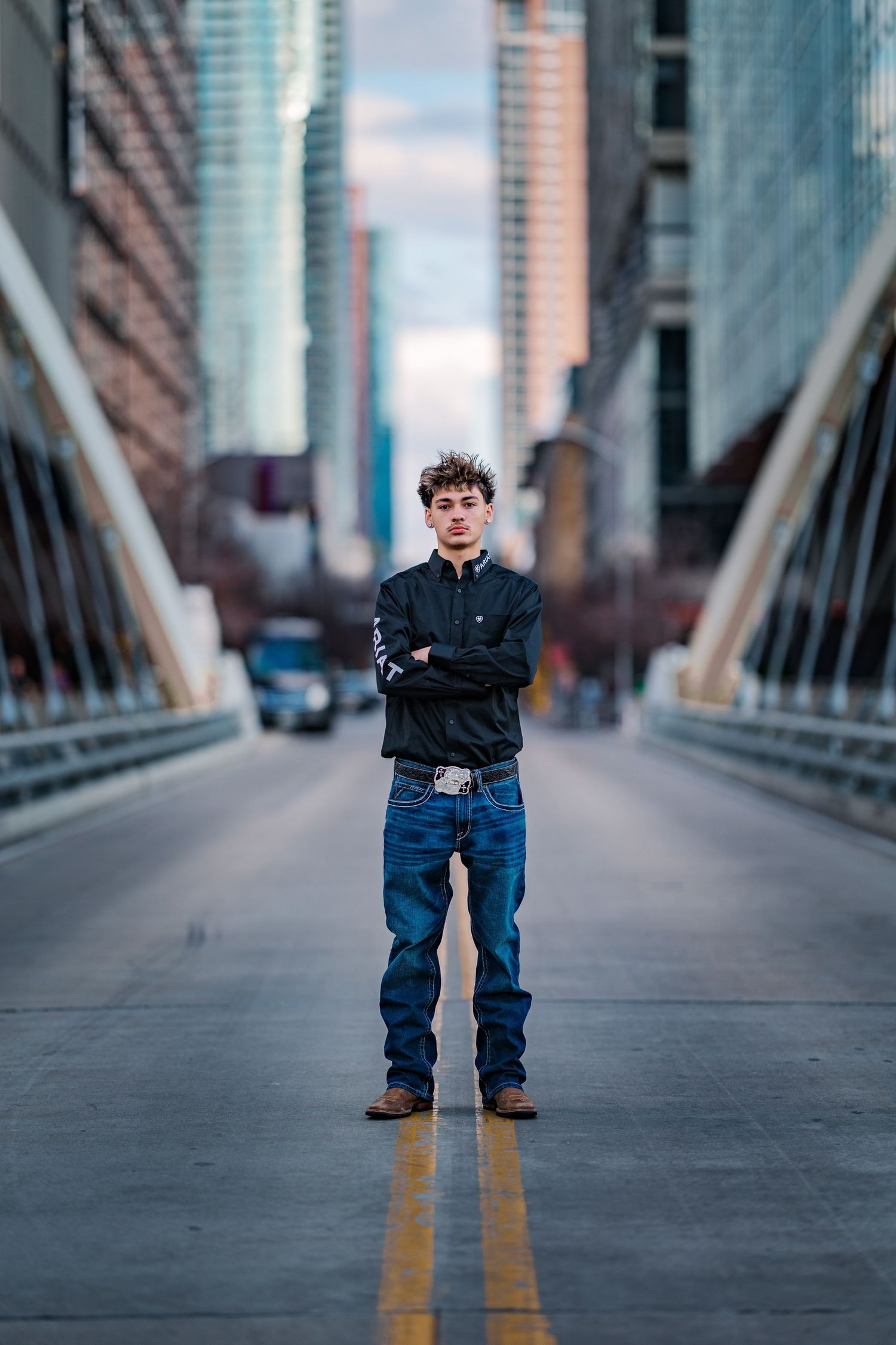 Confident young man standing arms crossed on city bridge with skyscrapers in background.