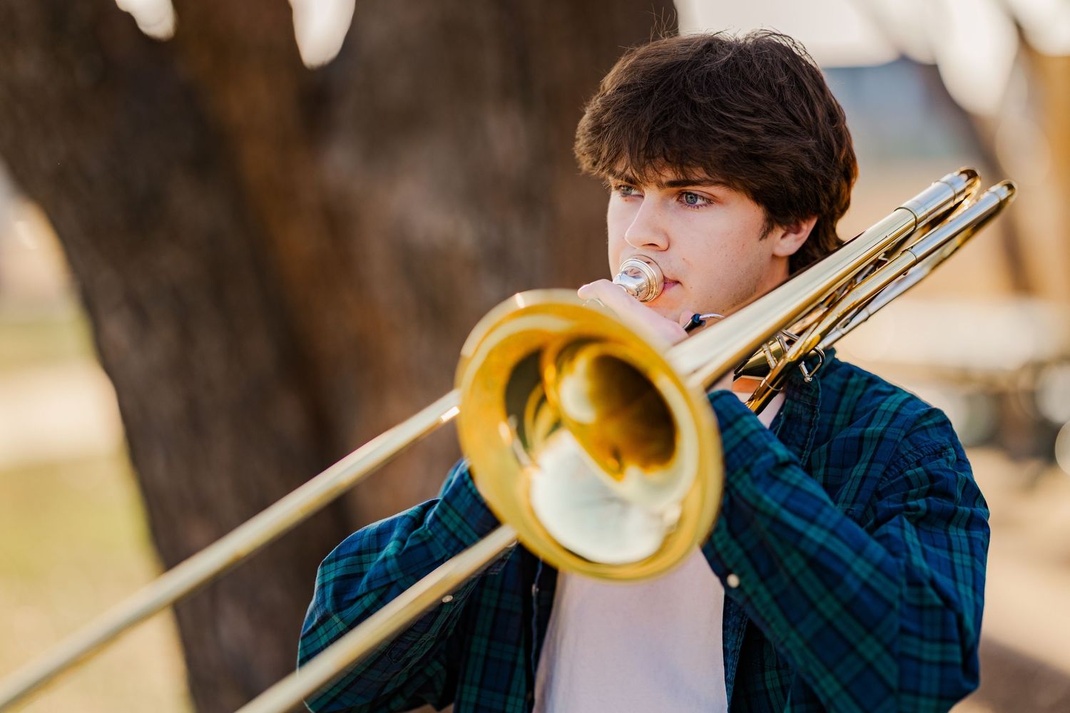 Teen boy plays trombone outdoors wearing a plaid shirt with blurred trees in the background.