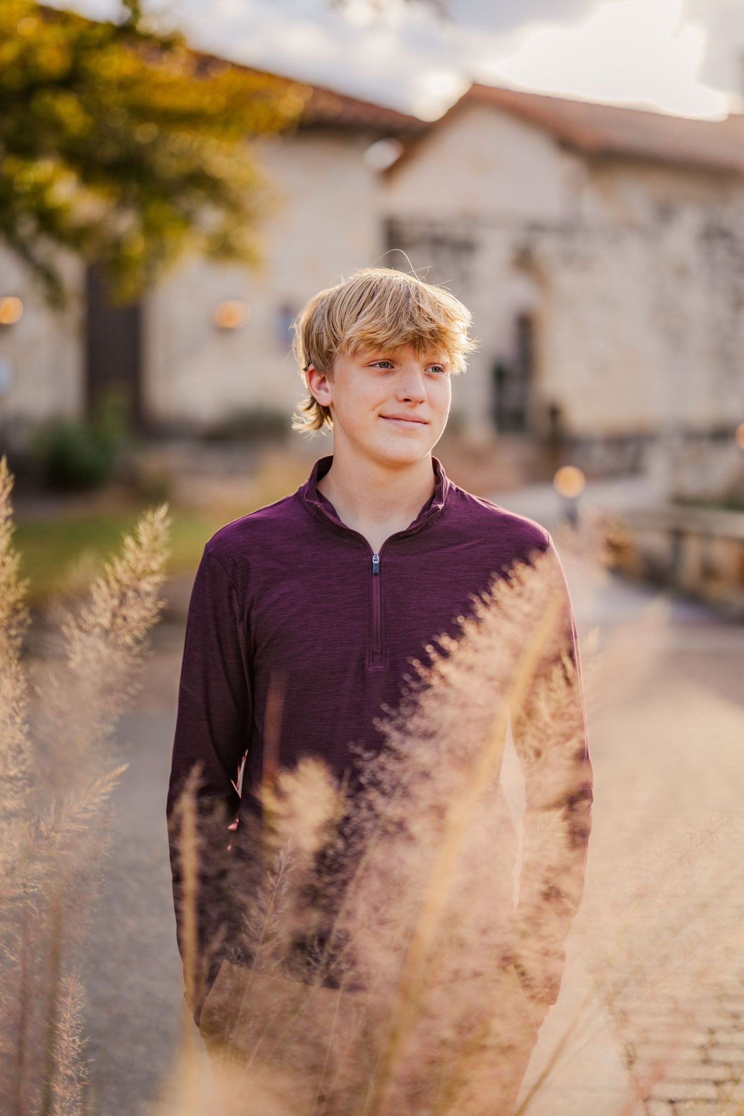 Blond teenage boy in purple pullover standing on brick path at golden hour.