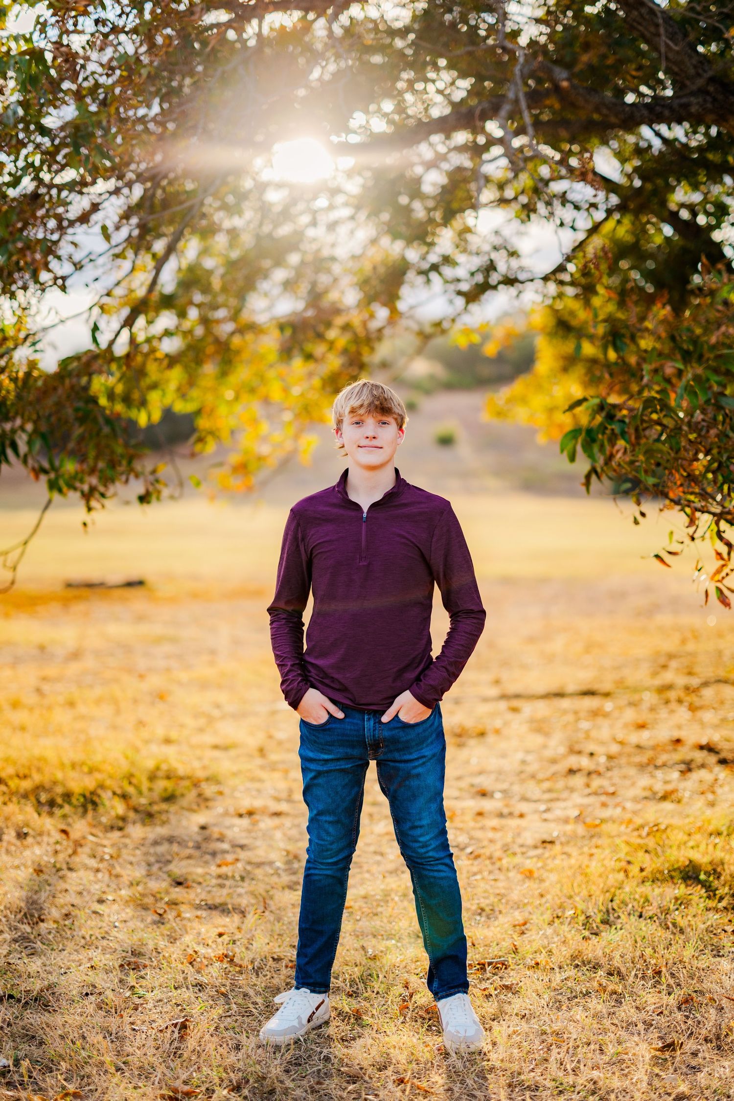 Teenage boy in purple polo and jeans stands under tree branches in golden autumn sunlit field.
