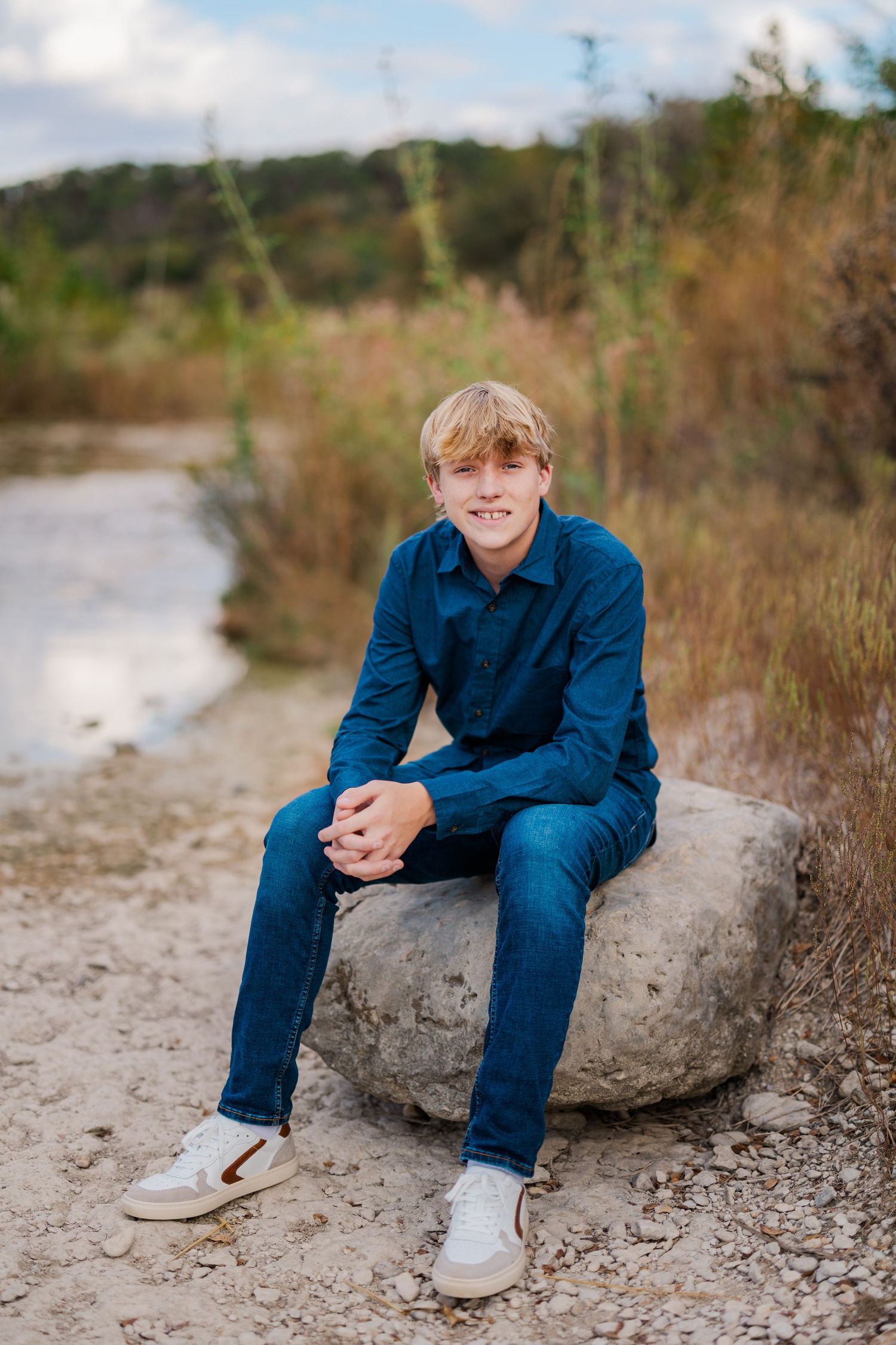 Smiling blond teen boy in blue denim shirt sitting on rock in natural outdoor setting.