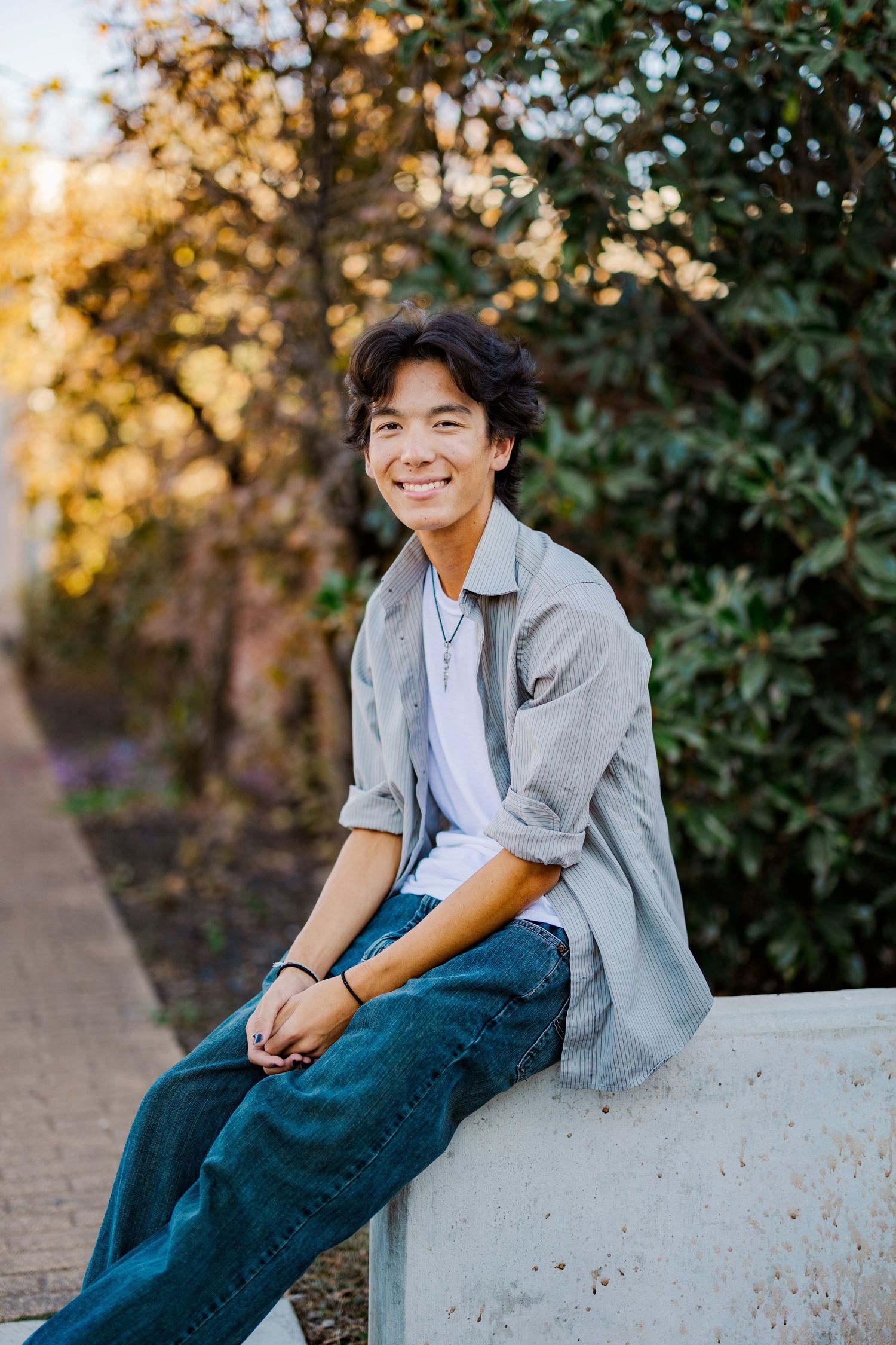 Smiling Asian teen boy in grey shirt and jeans sits on concrete wall surrounded by lush greenery.