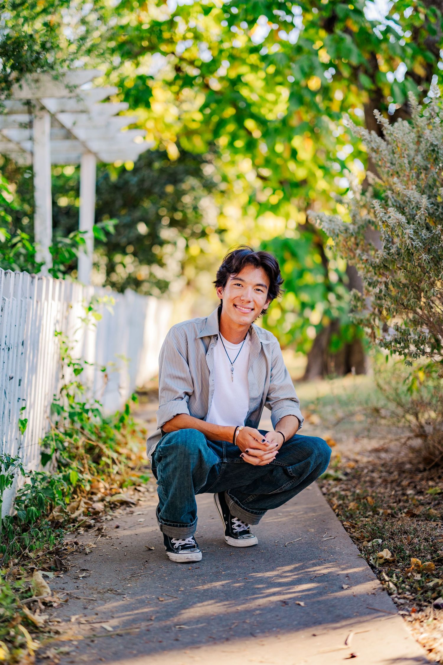 Smiling young man crouching on a garden path surrounded by lush green trees and a white fence.