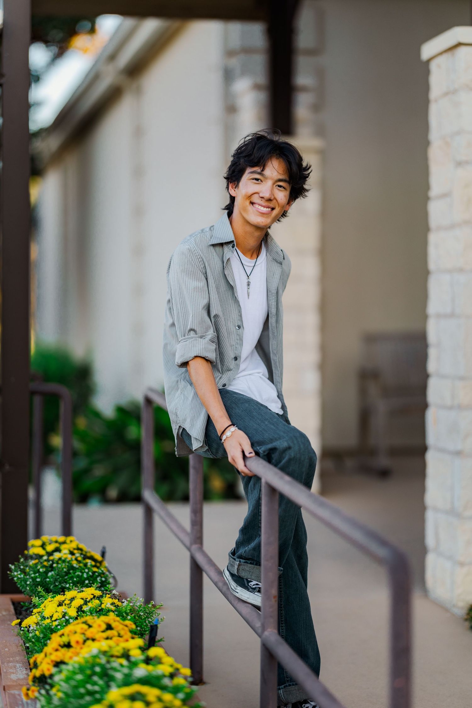 Smiling Asian teen boy sits casually on a railing beside yellow flowers outside a brick building.
