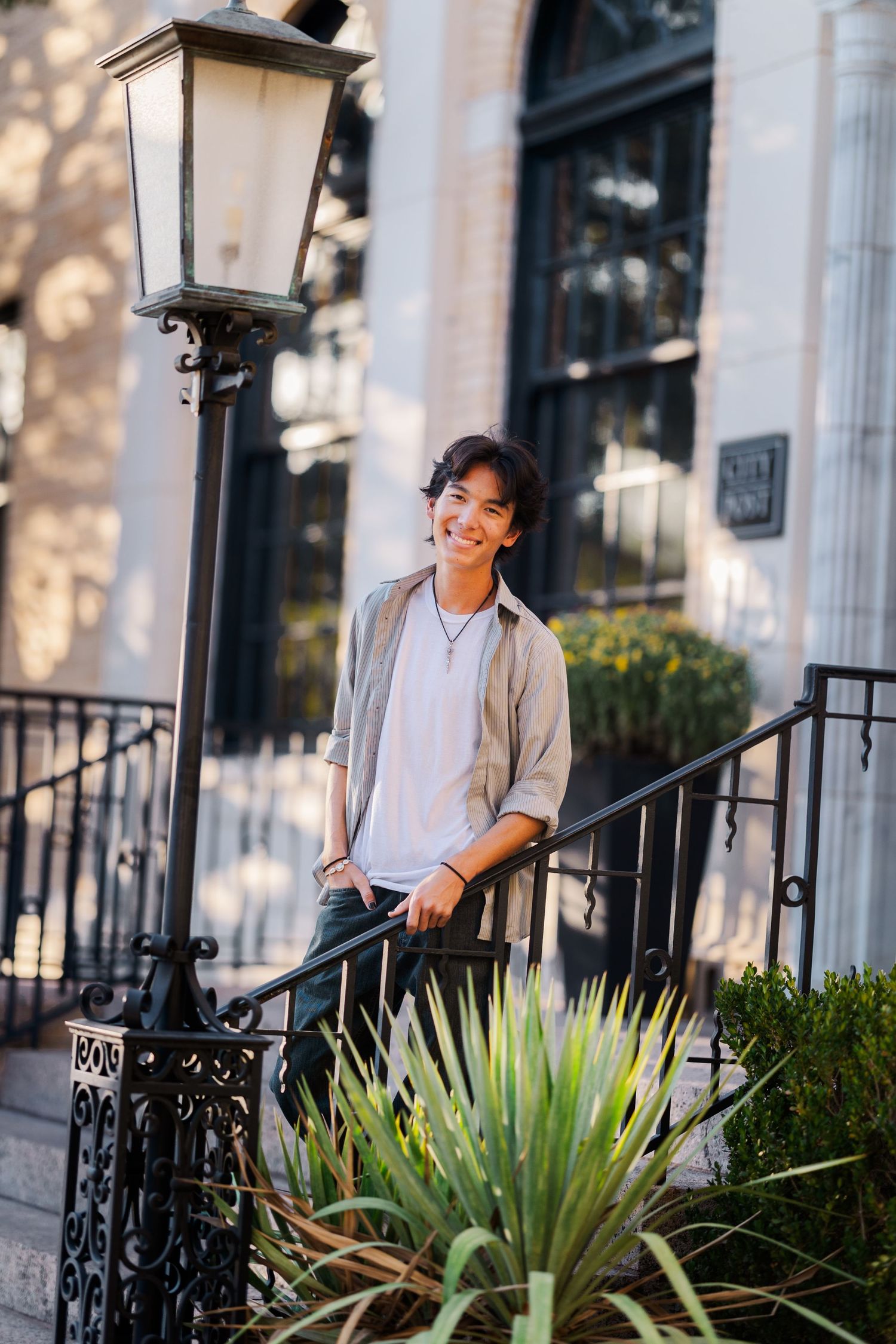 Smiling young man leaning on iron railing by a lamp post on sunny urban steps.
