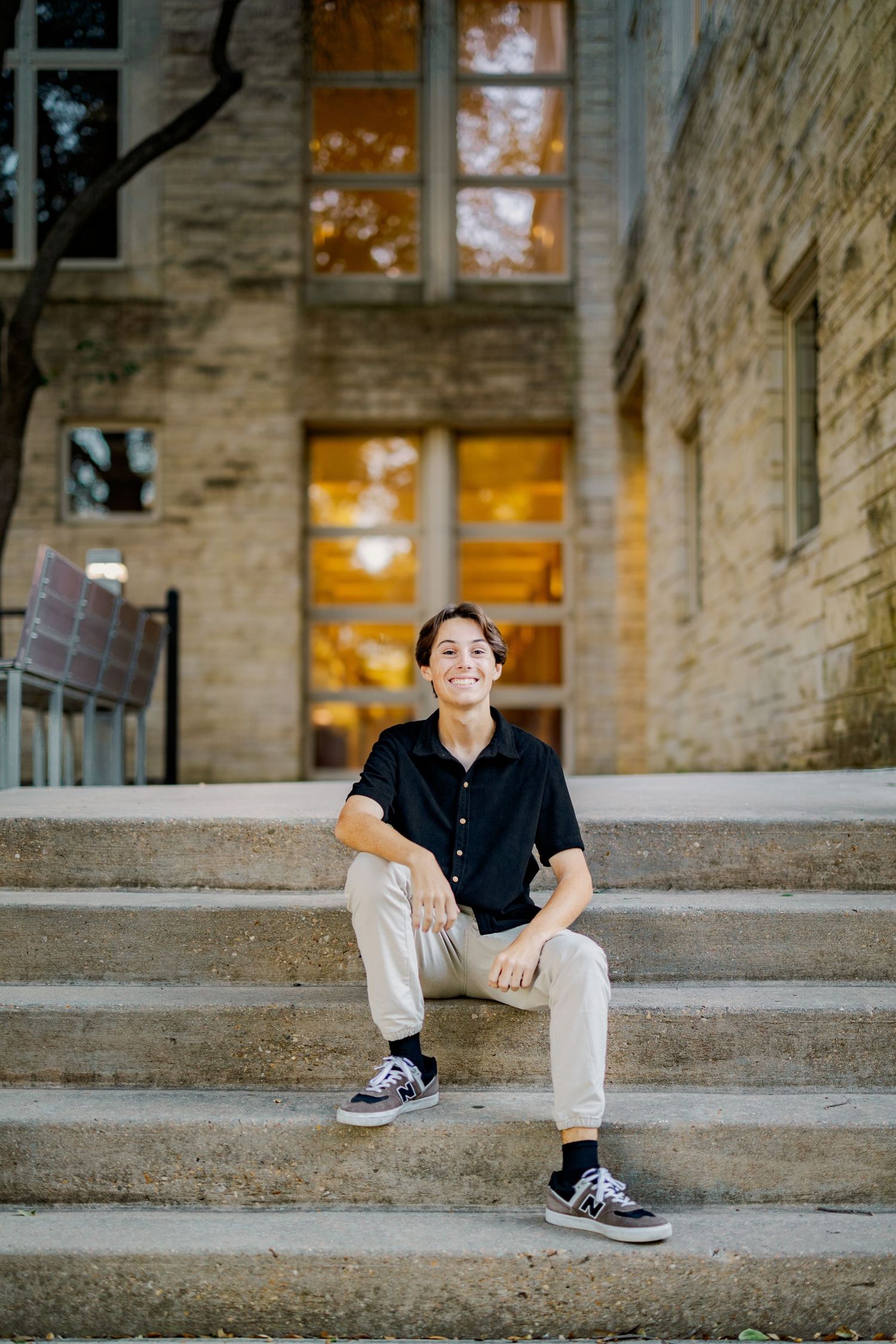 Smiling teen in black polo sits on stone steps outside a warmly lit historic building at dusk.