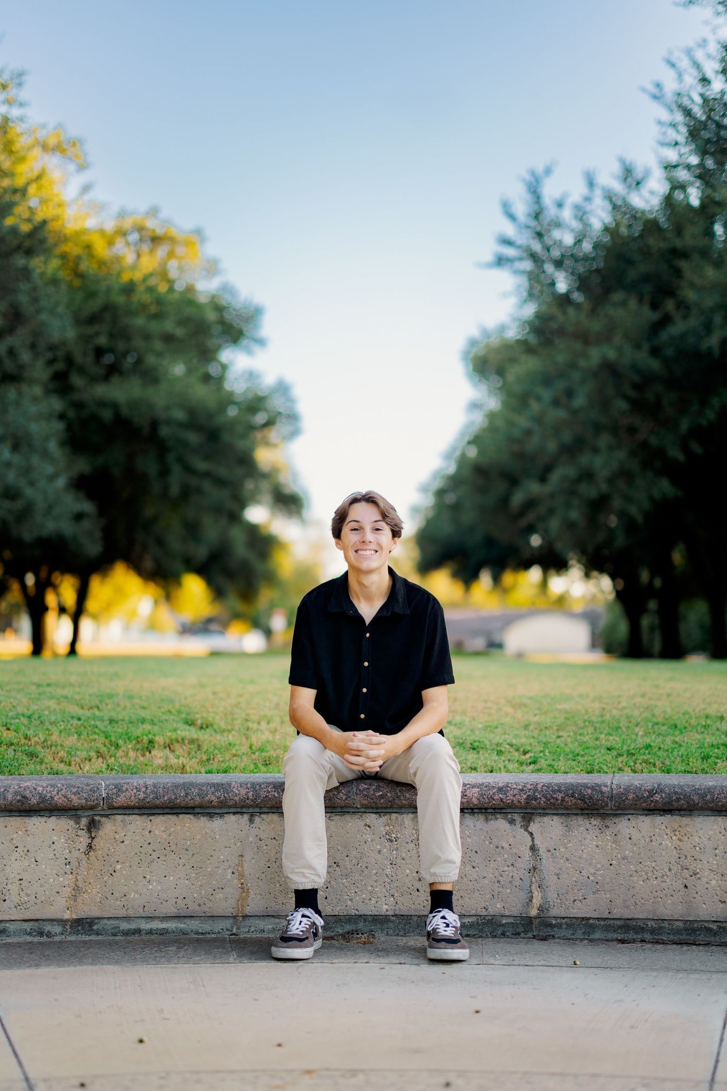 Smiling teen boy in black shirt sitting on stone ledge in tree-lined park.