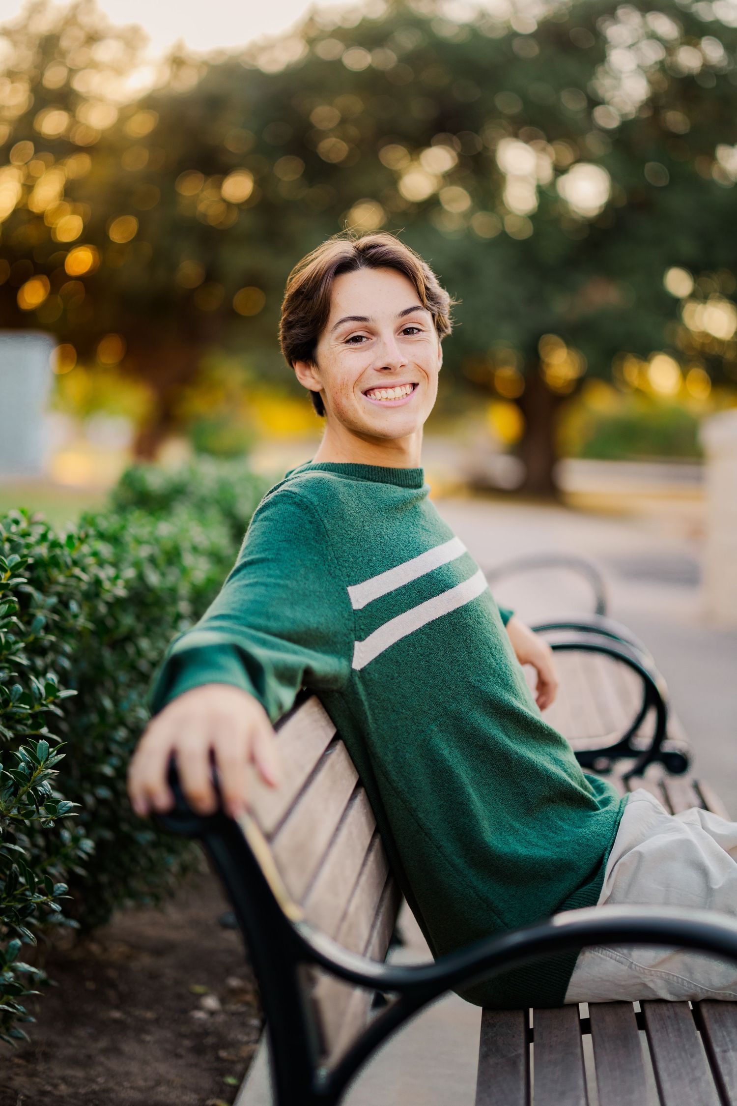 Smiling teen boy in green striped sweater sitting relaxed on a park bench at sunset.