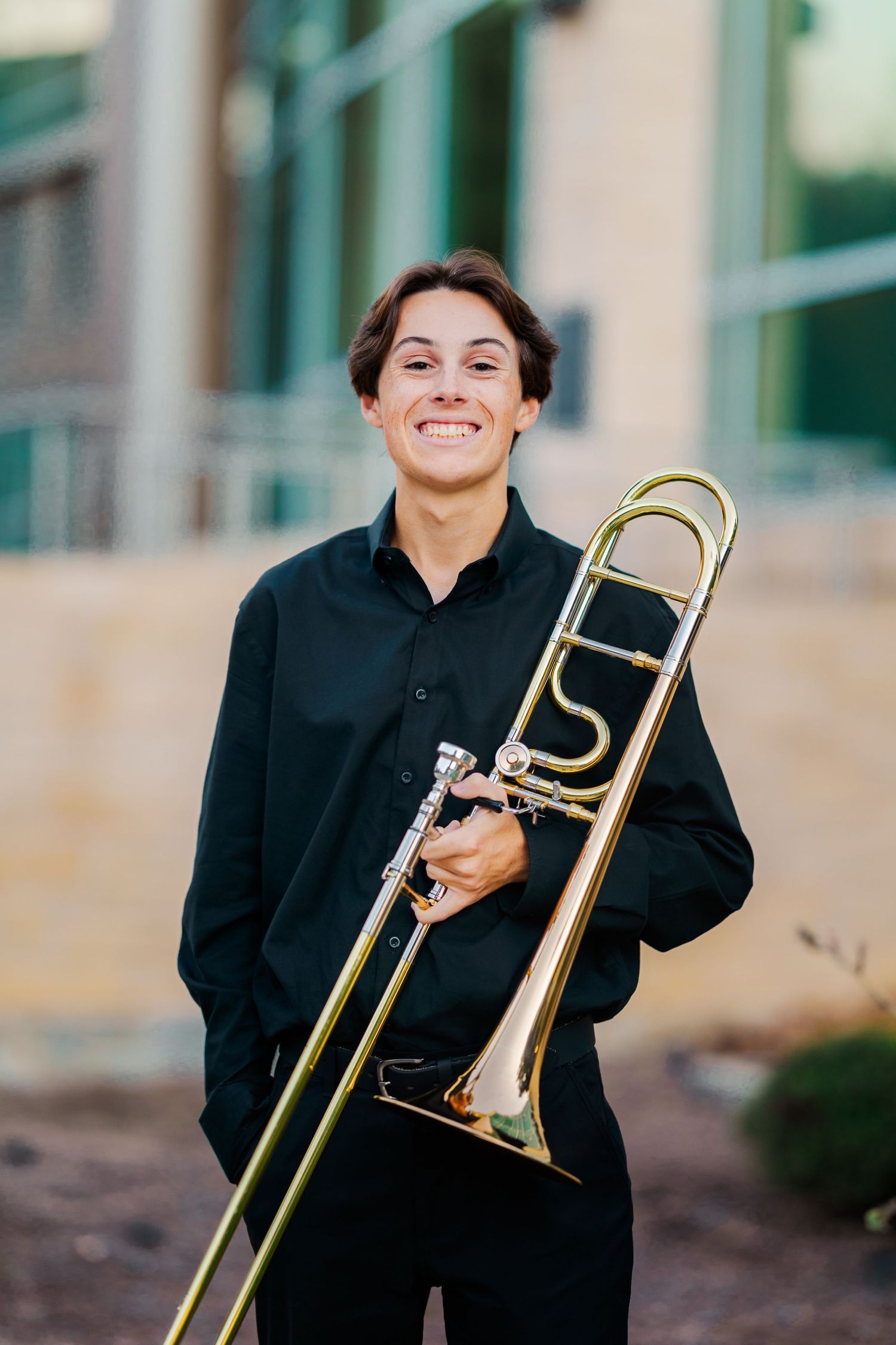 Smiling teenage boy in black shirt holding trombone for outdoor senior portrait near building.