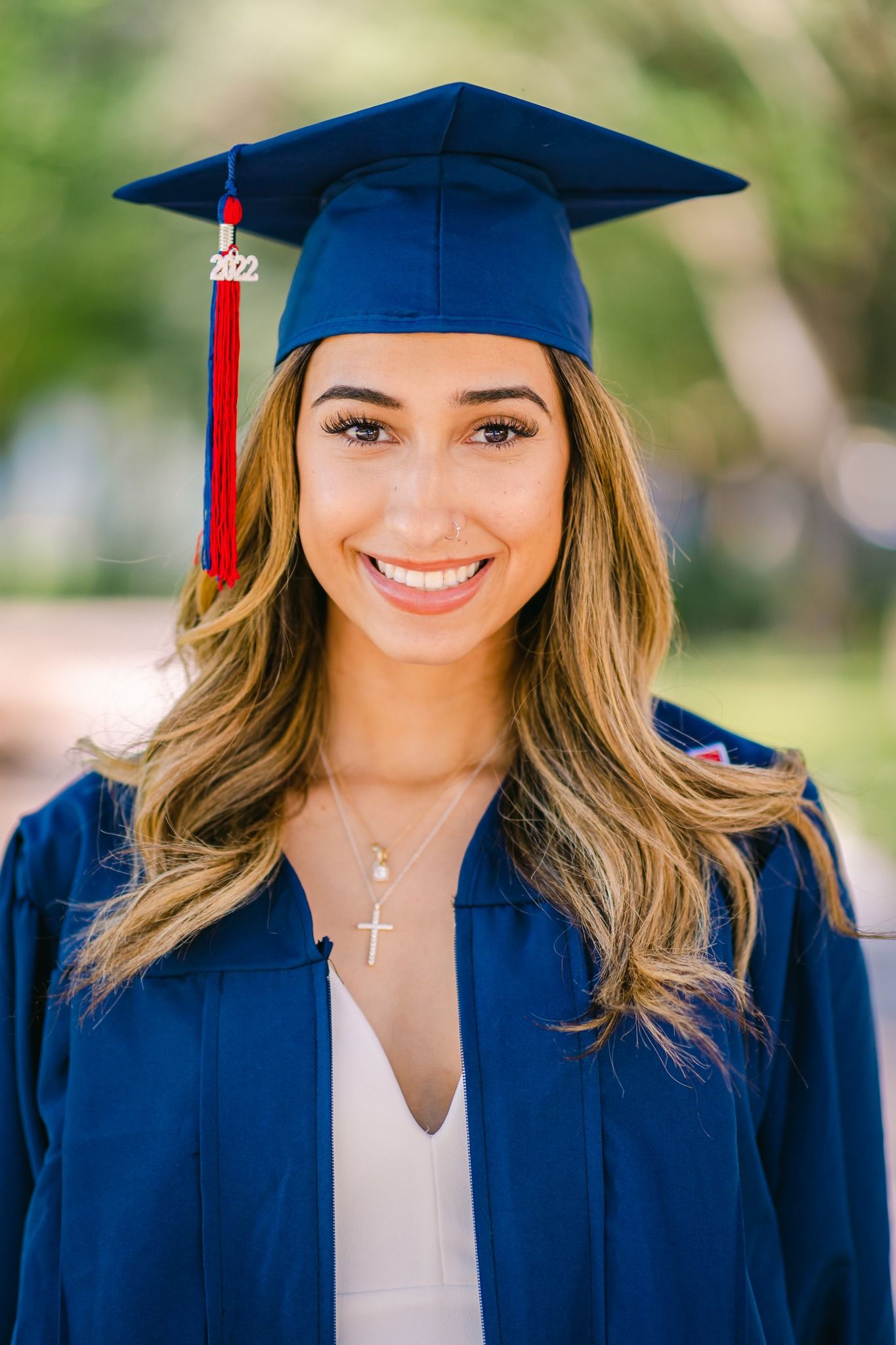 Smiling young woman in blue graduation cap and gown with red tassel and cross necklace outdoors.
