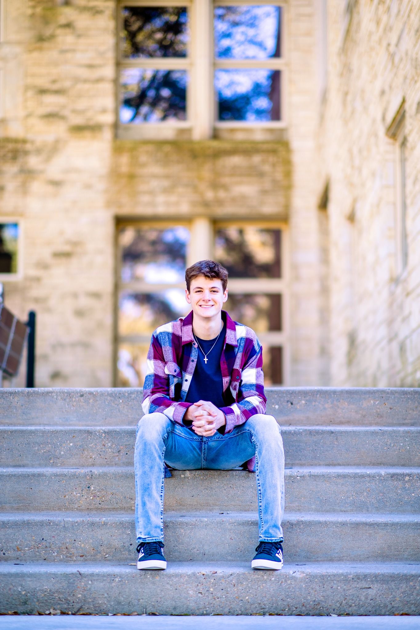 Smiling teenage boy in plaid shirt and jeans sits on stone steps outside a brick building.