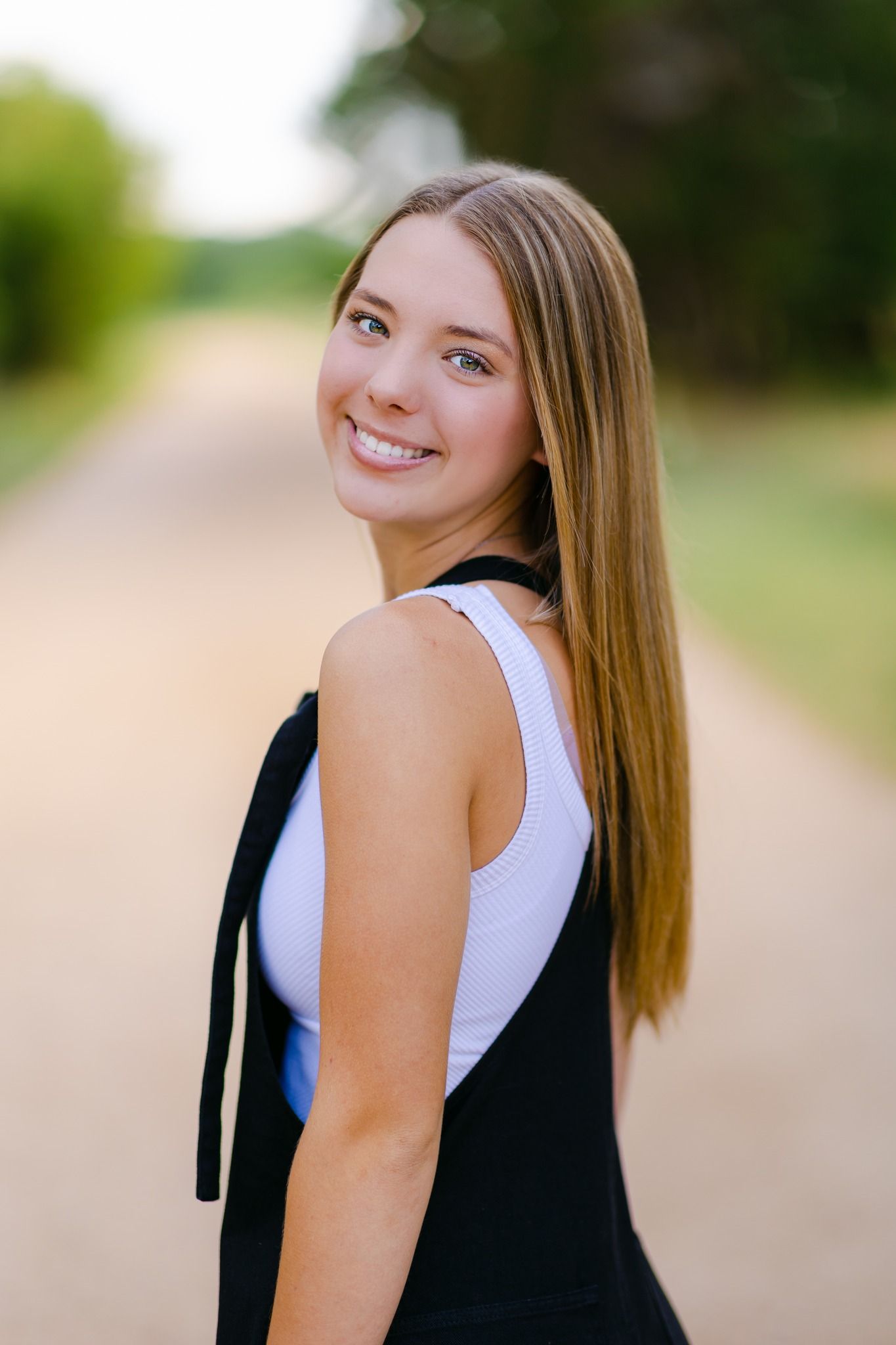 Smiling teenage girl with long blonde hair poses on a rural dirt road in a black and white outfit.