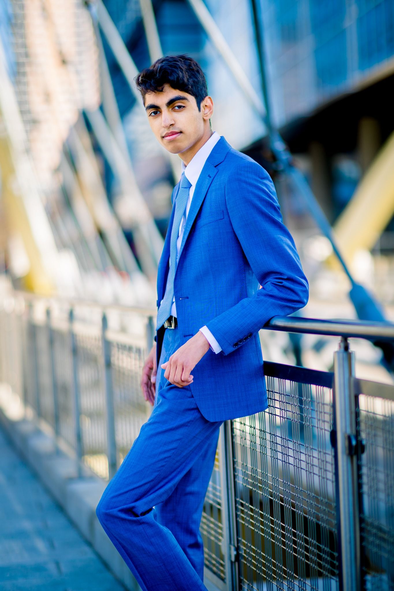 Confident young man in a royal blue suit leans against a railing in a modern urban setting.