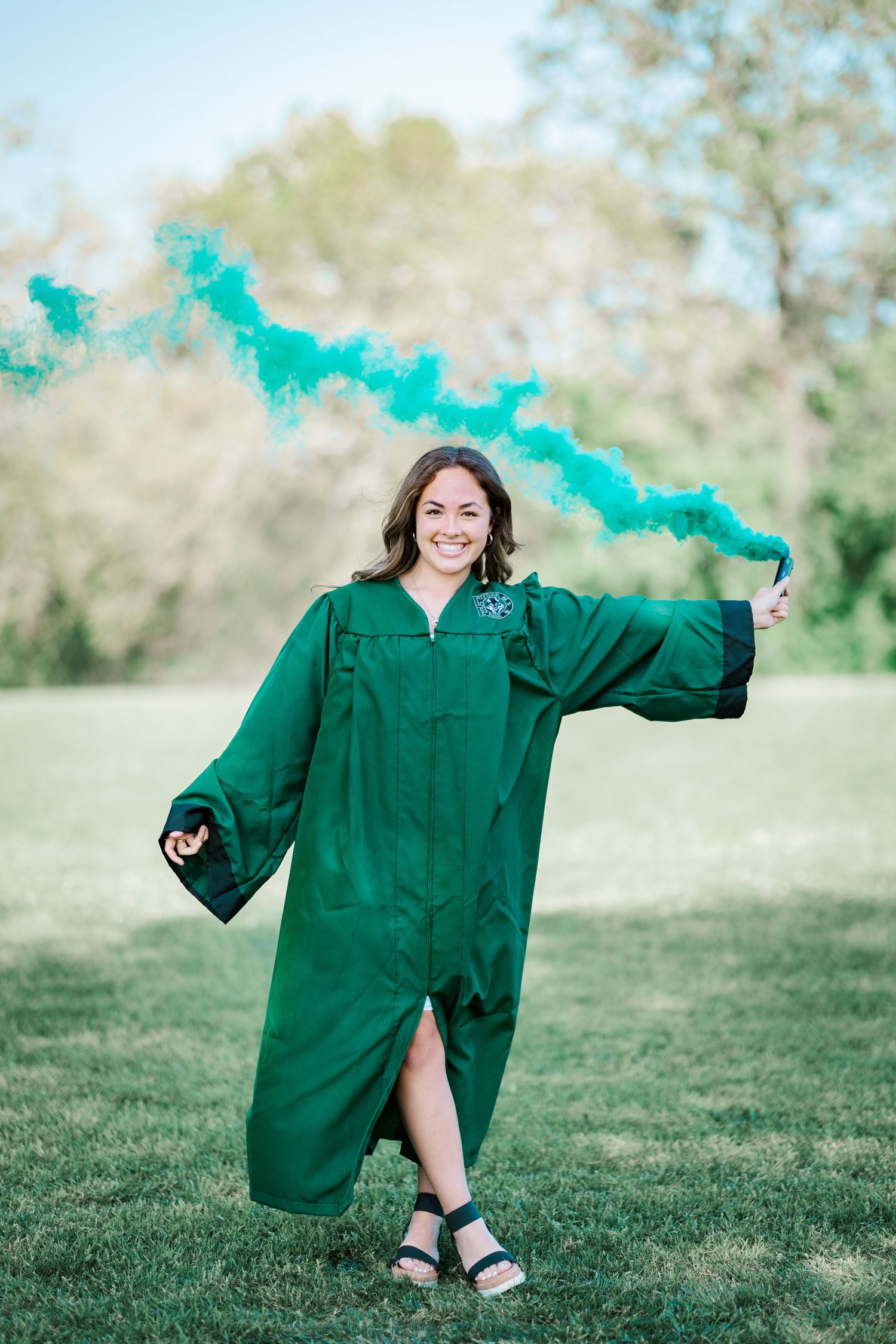 Smiling graduate in green cap and gown holding teal smoke flare outdoors on grass.