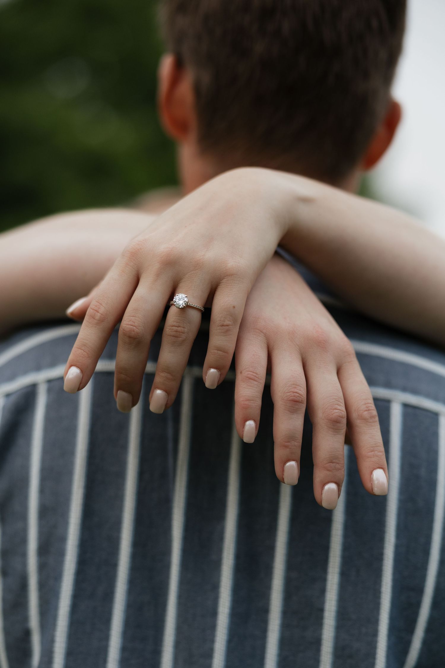 Close-up of hands with light pink nail polish and engagement ring resting on a striped chair backrest.