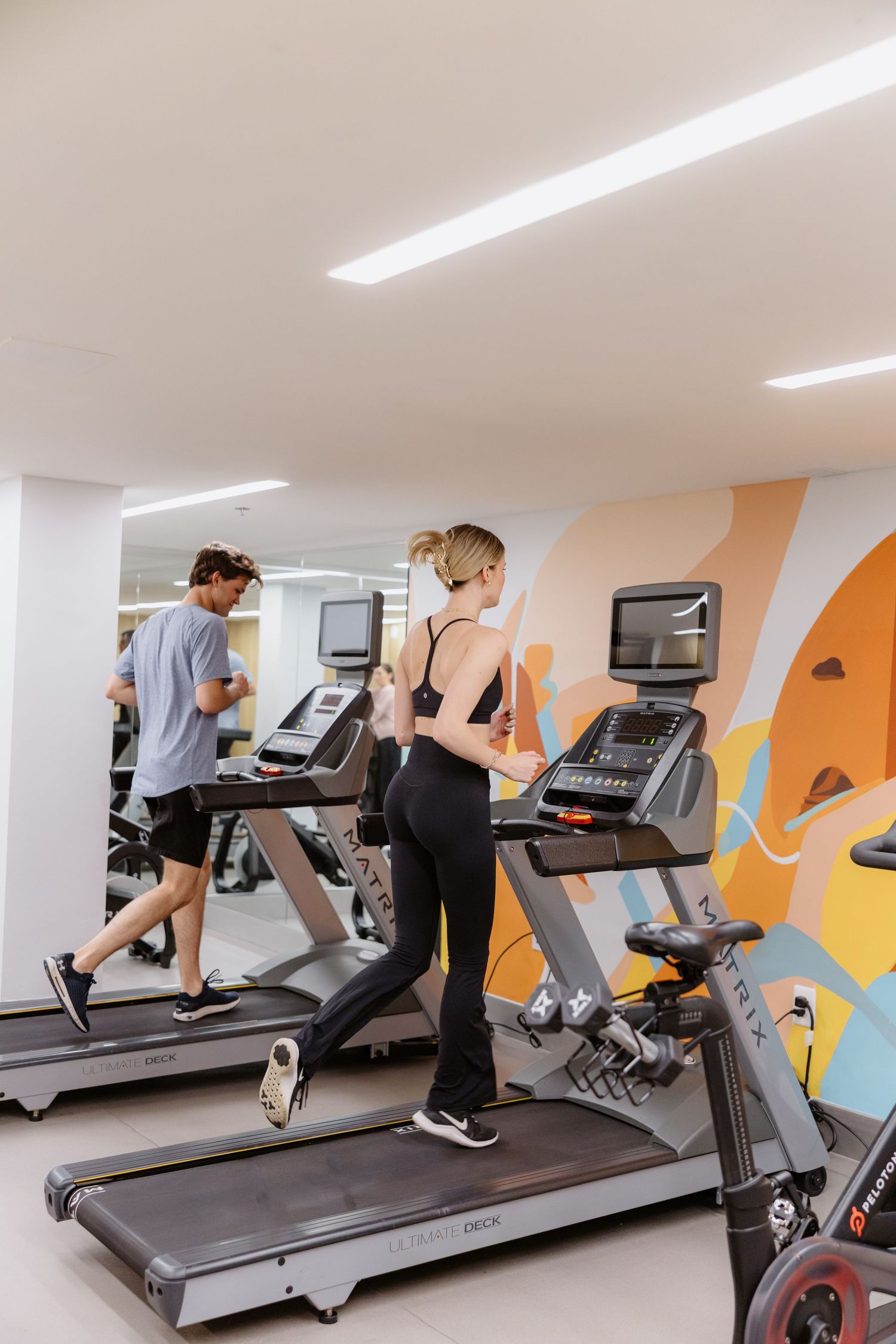People exercising on treadmills in a modern gym with colorful mural wall art and bright lighting.