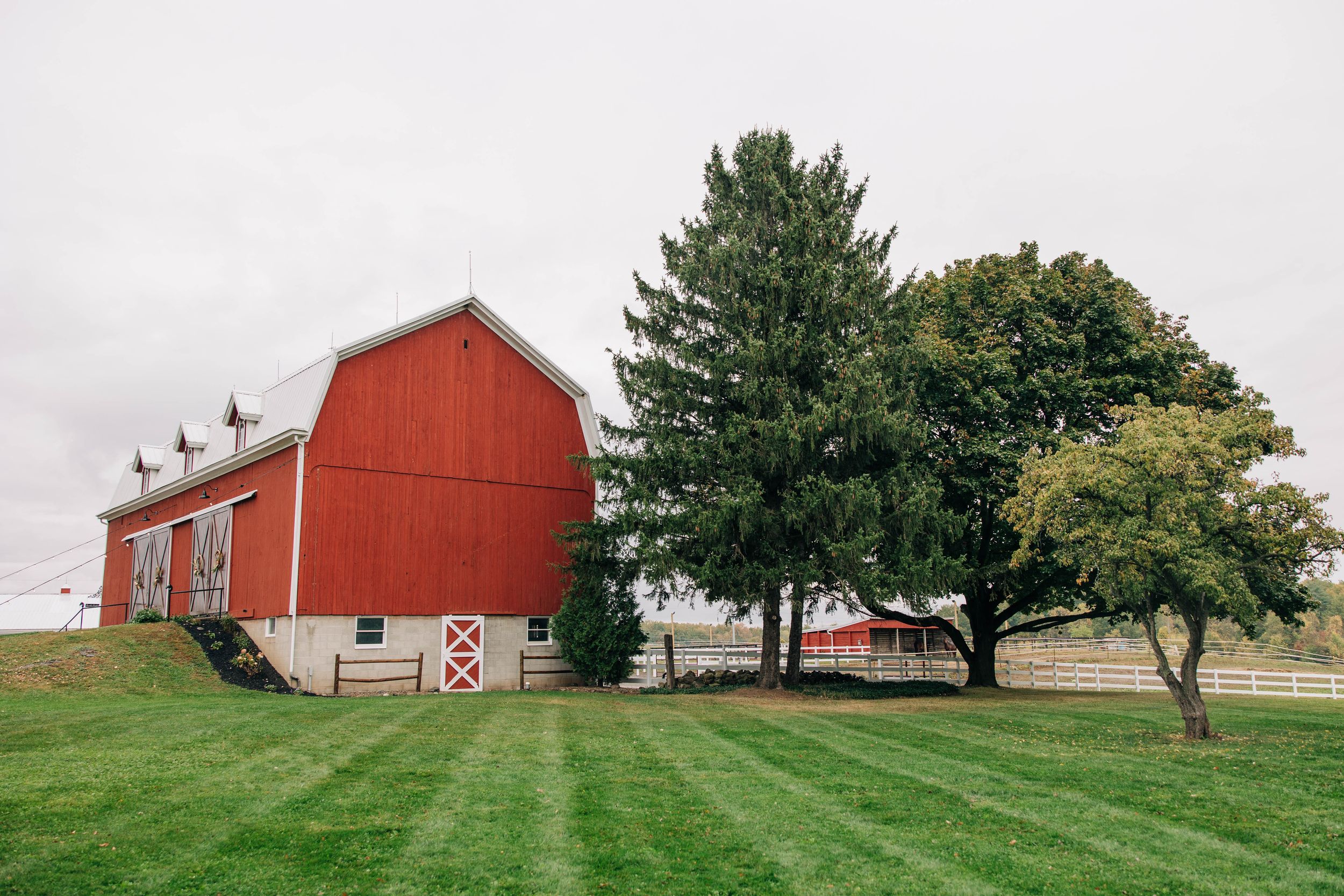 A red barn stands on a green lawn with mature trees and a cloudy sky in the background.