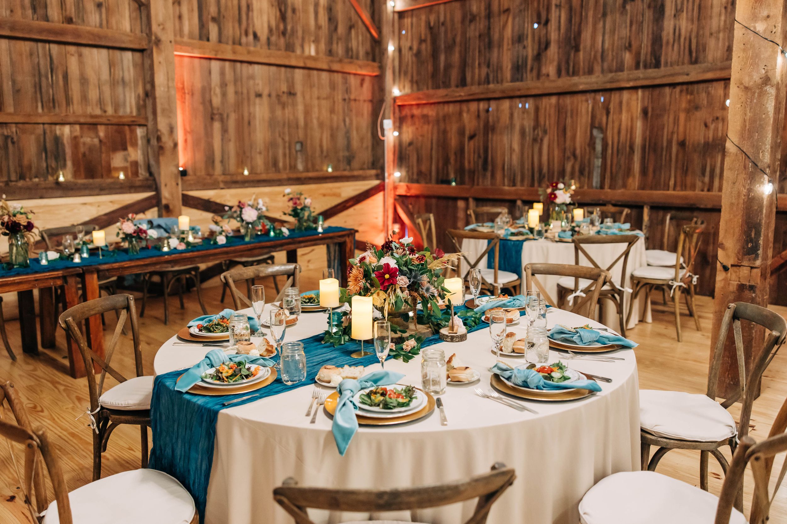 Rustic barn wedding reception table set with teal blue linens, white tablecloths and wooden cross-back chairs.