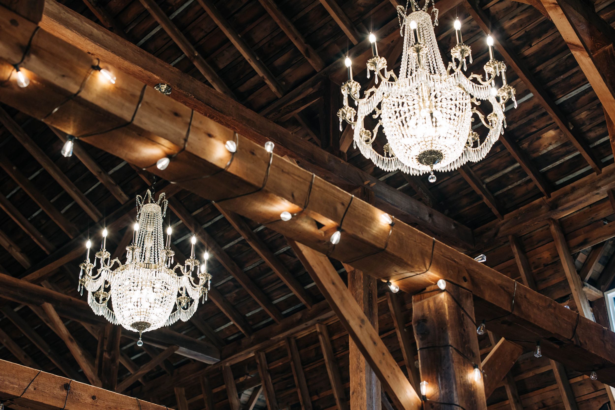 Crystal chandeliers hang from a rustic wooden beam ceiling in an elegant barn venue interior.