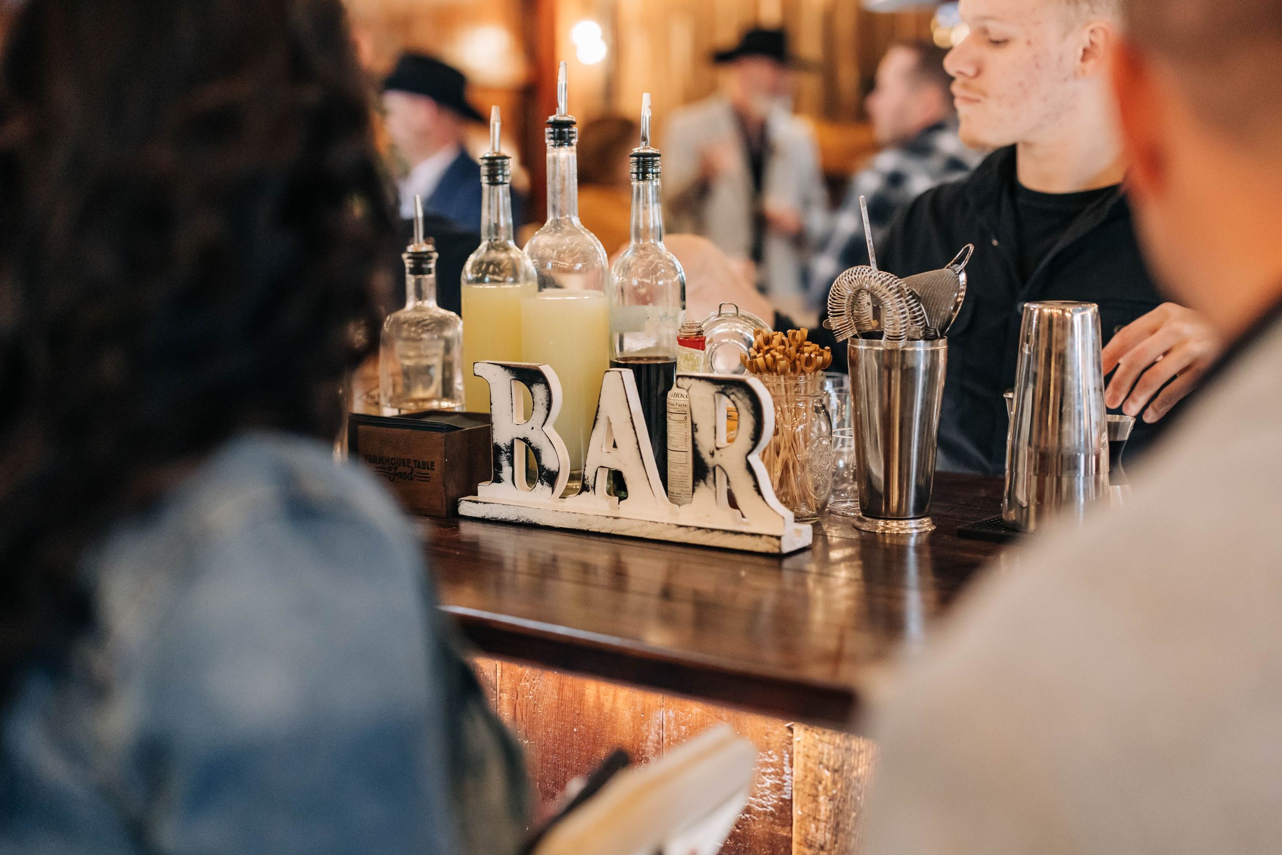 A wooden bar sign and assorted liquor bottles on display at a cozy bar counter during an evening gathering.