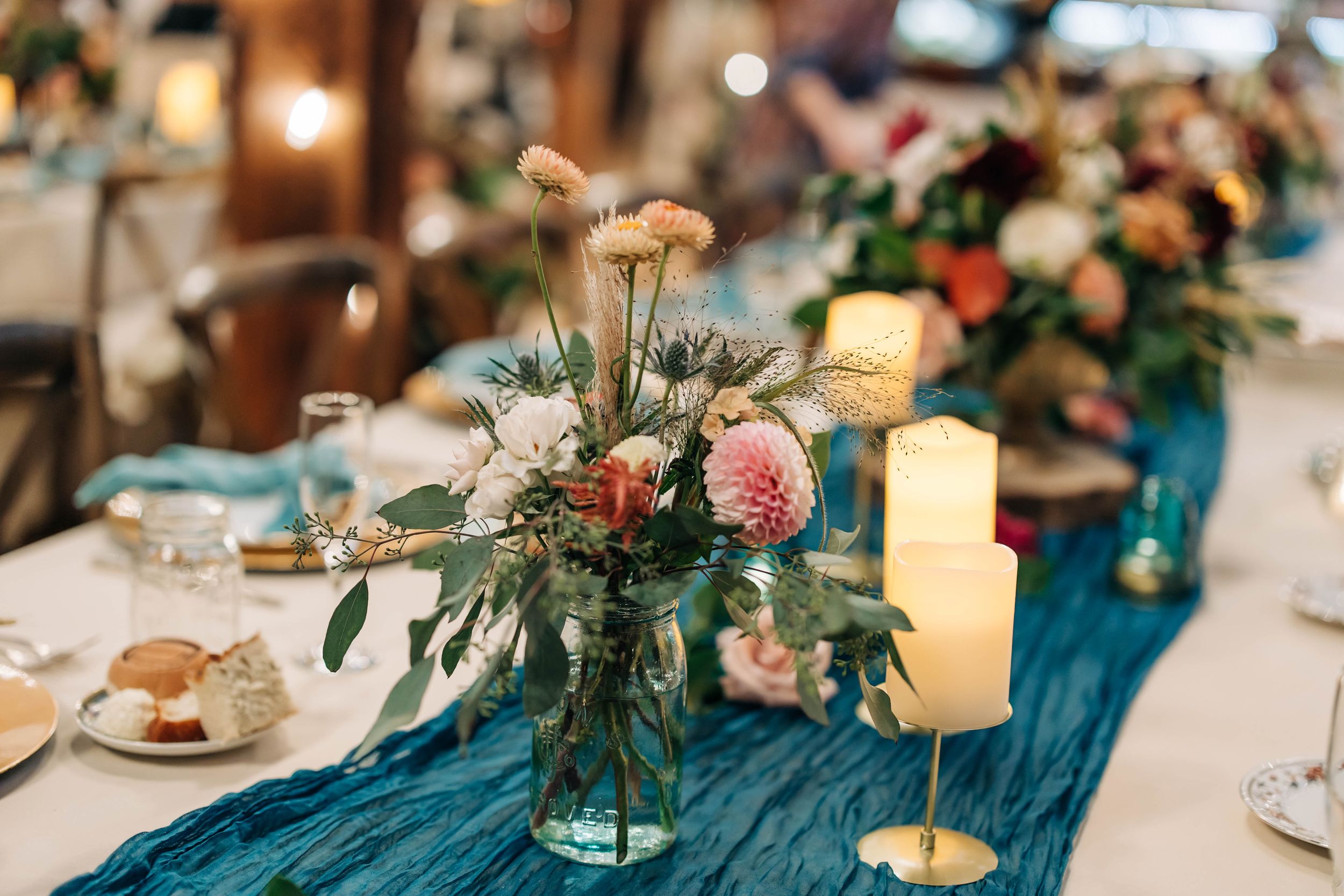 Elegant wedding reception table with teal blue runner, floral centerpieces and glowing candles in a rustic venue setting.