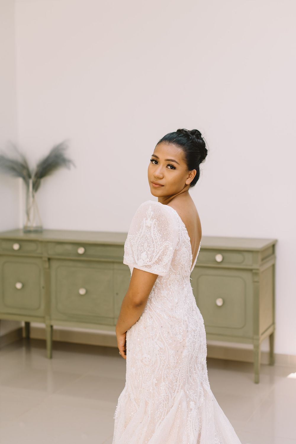 Woman in a white lace wedding dress poses against a light backdrop with green vintage dressers.