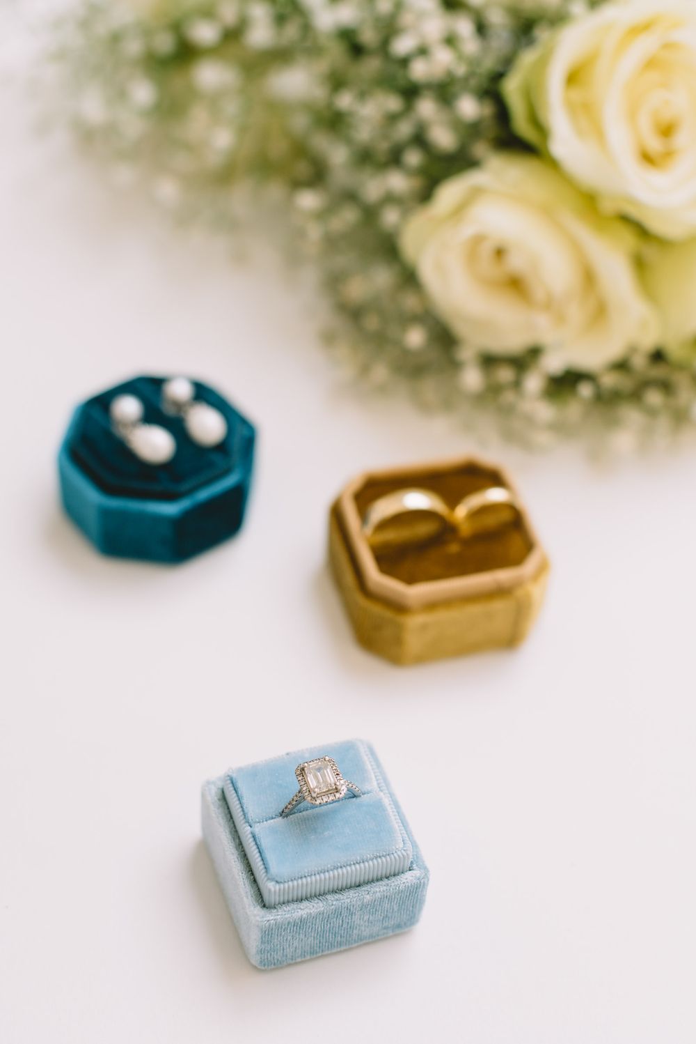 Stylish engagement ring boxes in turquoise, gold, and light blue velvet displayed with white roses and baby's breath flowers.