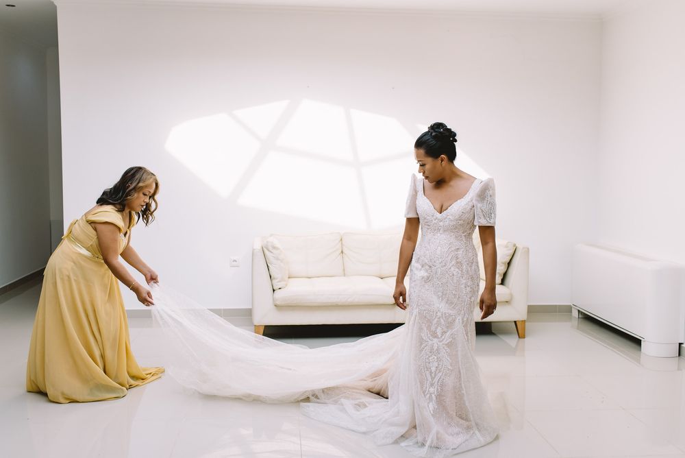 Two people in elegant dresses in a bright white room, one helping adjust a long flowing wedding gown train.