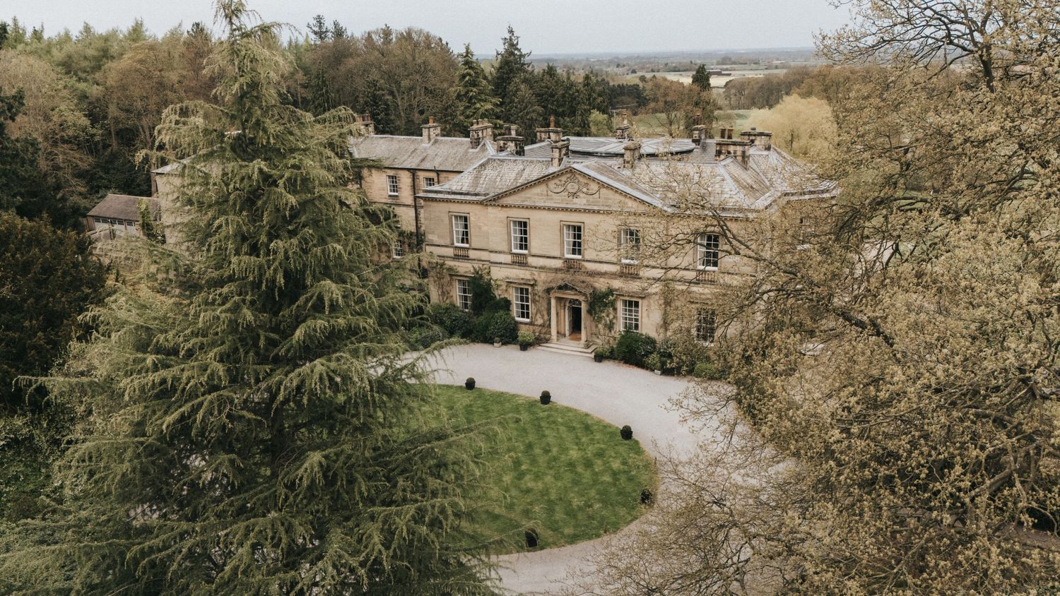 Elegant historic stone mansion with circular driveway nestled among trees in a countryside setting.