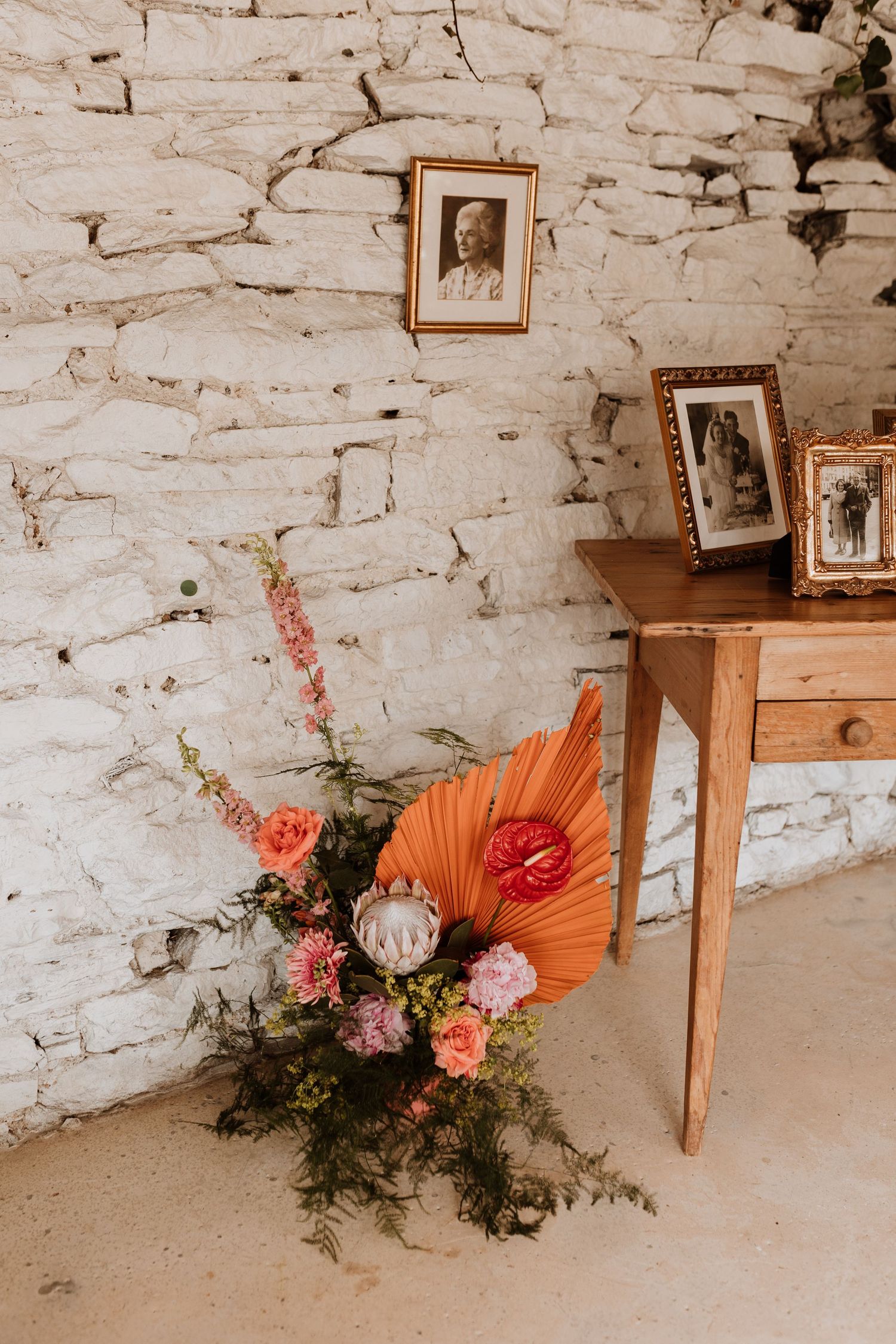 Fun dried palm fronds and fresh floral arrangements decorate a rustic whitewashed stone wall with vintage framed photos.