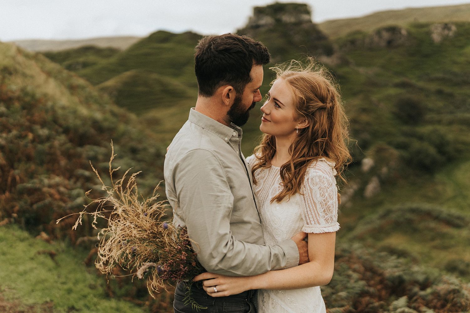 A couple embraces in a romantic moment against a backdrop of rolling green hills and rural landscape in Scotland.