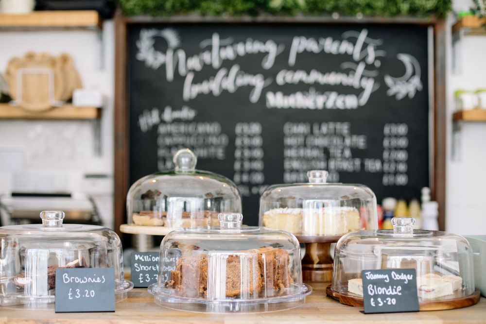 Glass cake stands with domed covers displayed in front of a chalkboard menu at a bakery counter.