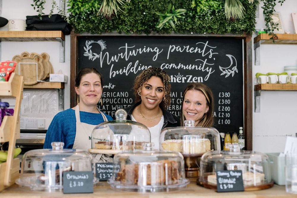Three people smiling behind a bakery counter with jars of goods and a chalkboard menu on display.