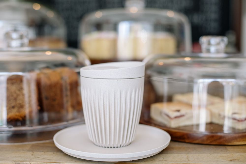 White ribbed ceramic coffee cup and saucer on a wooden surface with glass domed pastry displays in background.