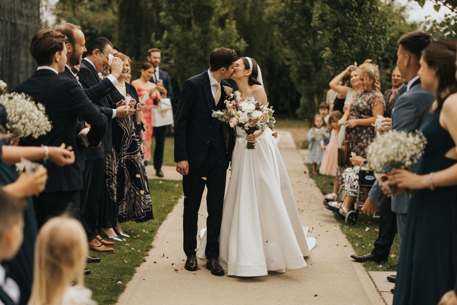 Wedding guests celebrate and throw confetti as newlyweds share their first kiss on a tree-lined outdoor path.