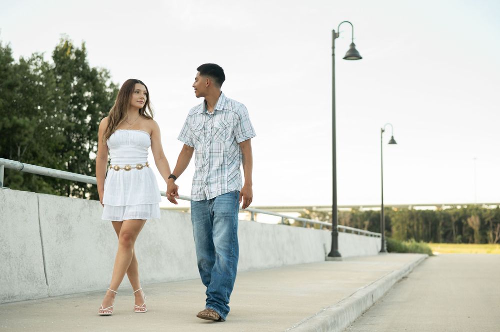 A couple in white outfits walks together along a concrete path during a romantic outdoor photoshoot.