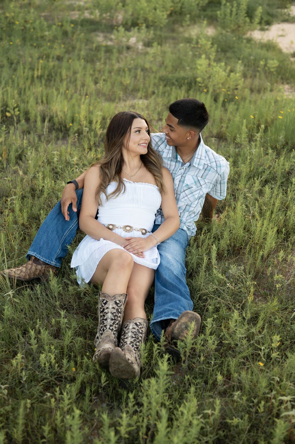 A couple sits intimately together in tall green grass while wearing denim and western boots in a rustic outdoor setting.