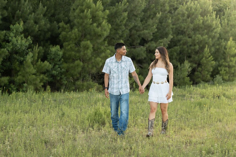 A romantic couple walking hand in hand through a summer meadow with evergreen trees in the background.