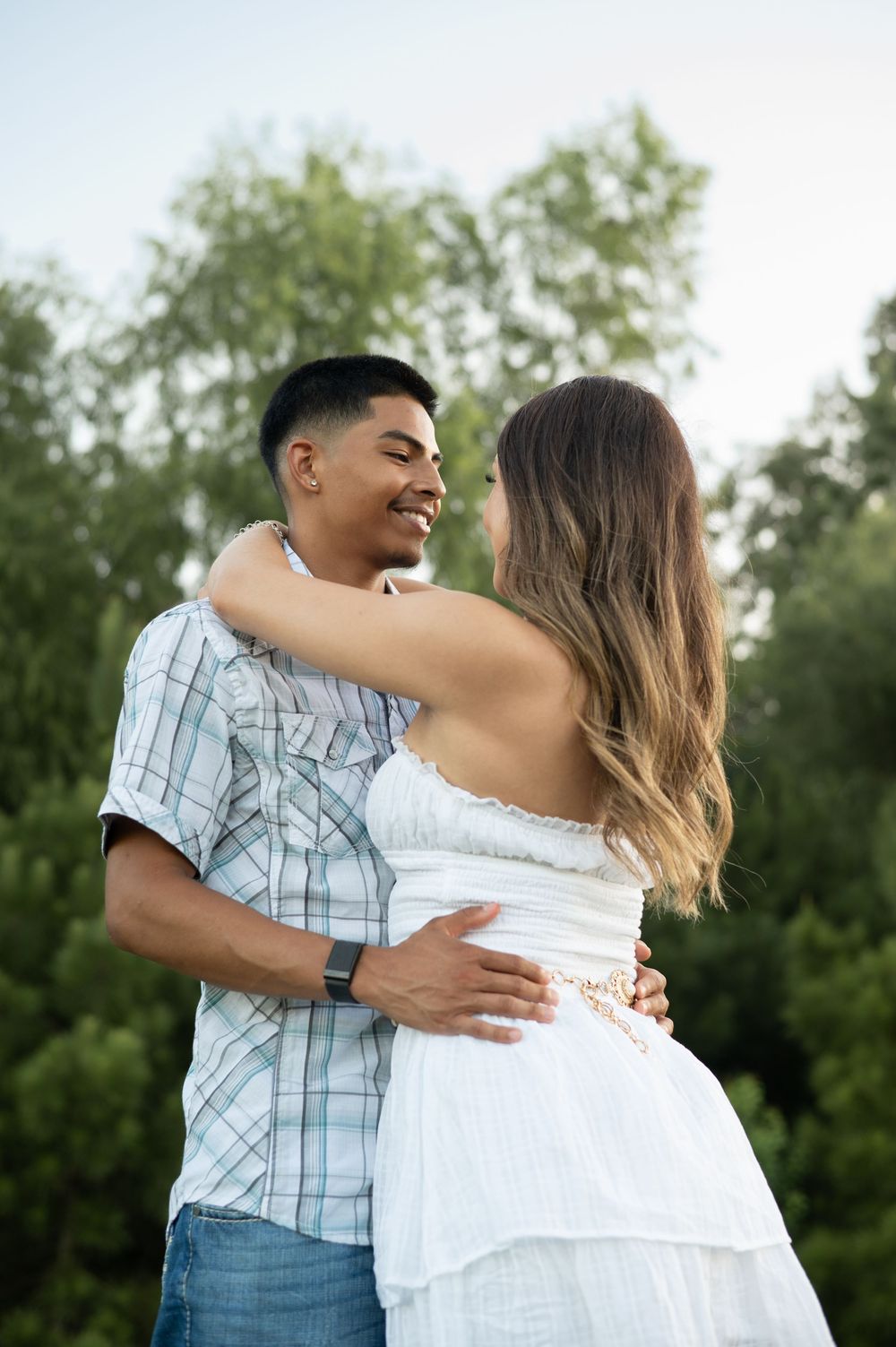 A romantic couple embraces outdoors in a sunny garden setting, with green trees in the background.
