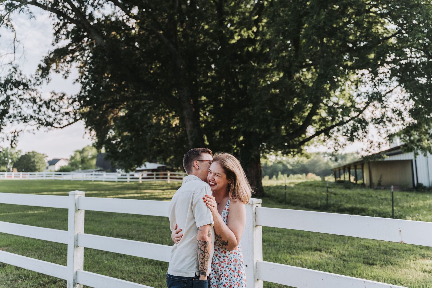 A couple shares a romantic moment by a white fence at sunset in an outdoor farm setting.