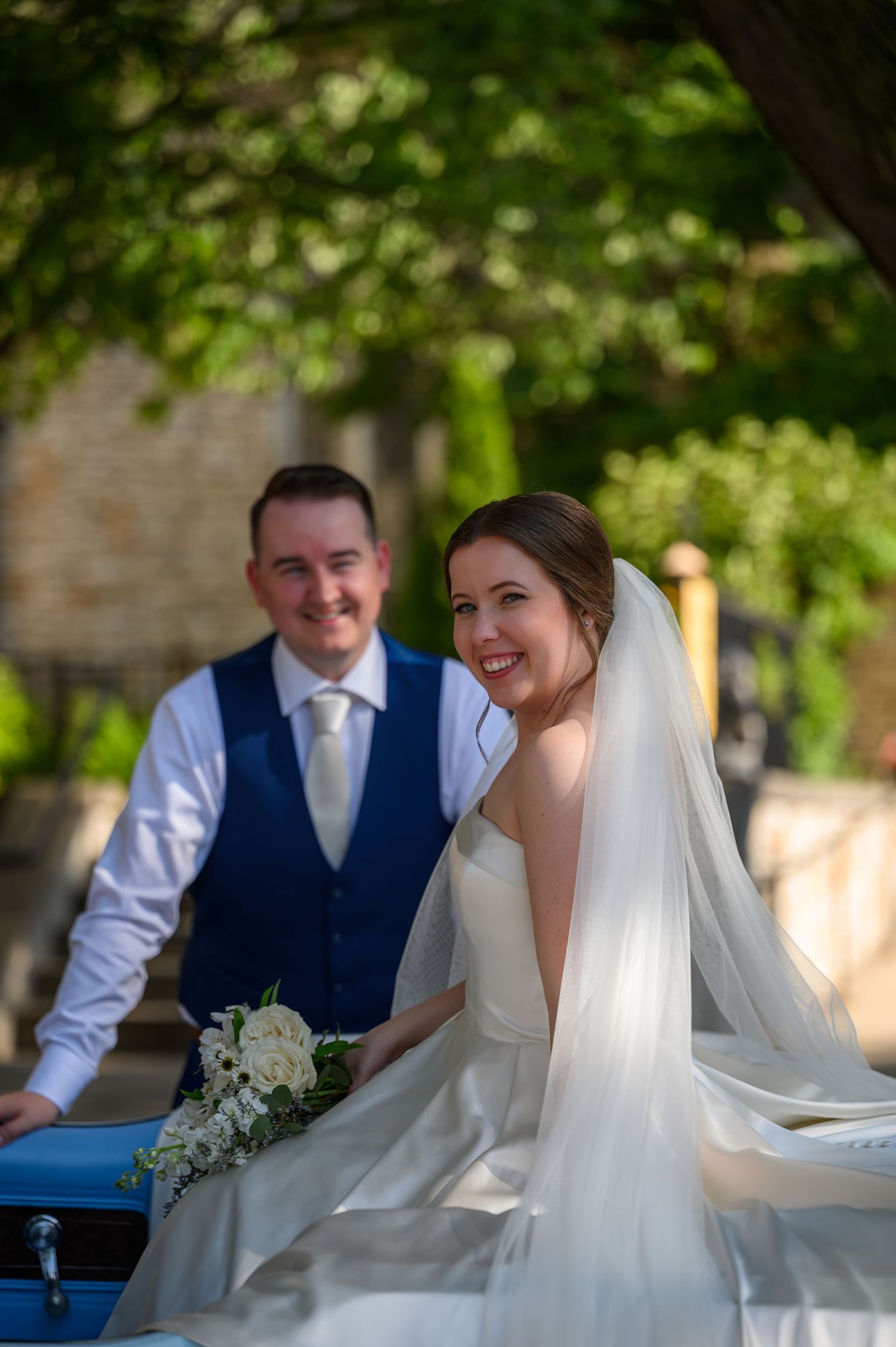 A happy bride in a flowing white dress and veil sits next to her groom in a navy vest on their wedding day.